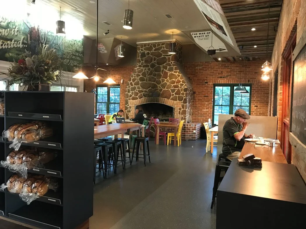 Inside the Trappers cafe. Visible is a brick and stone fireplace, colouful chairs, a large wooden table, and a man eating at a counter.