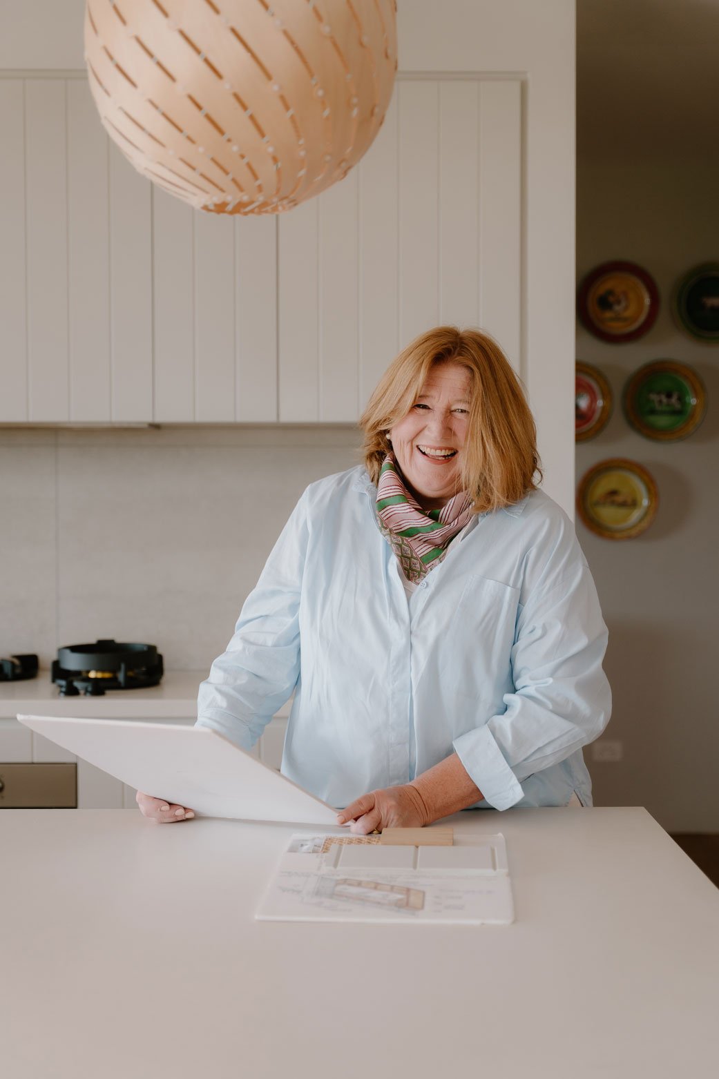 Suzie, wearing a colourful scarf smiling while holding a large white board in a modern kitchen.