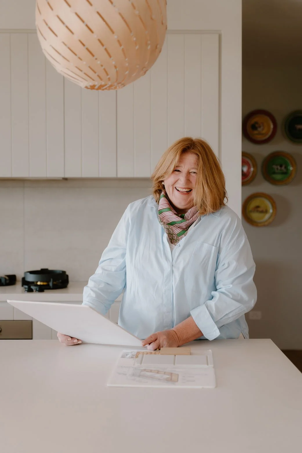 Suzie standing at a kitchen bench, holding a board. There are design plans on the bench. Suzie is looking towards the camera, with a big smile on her face.