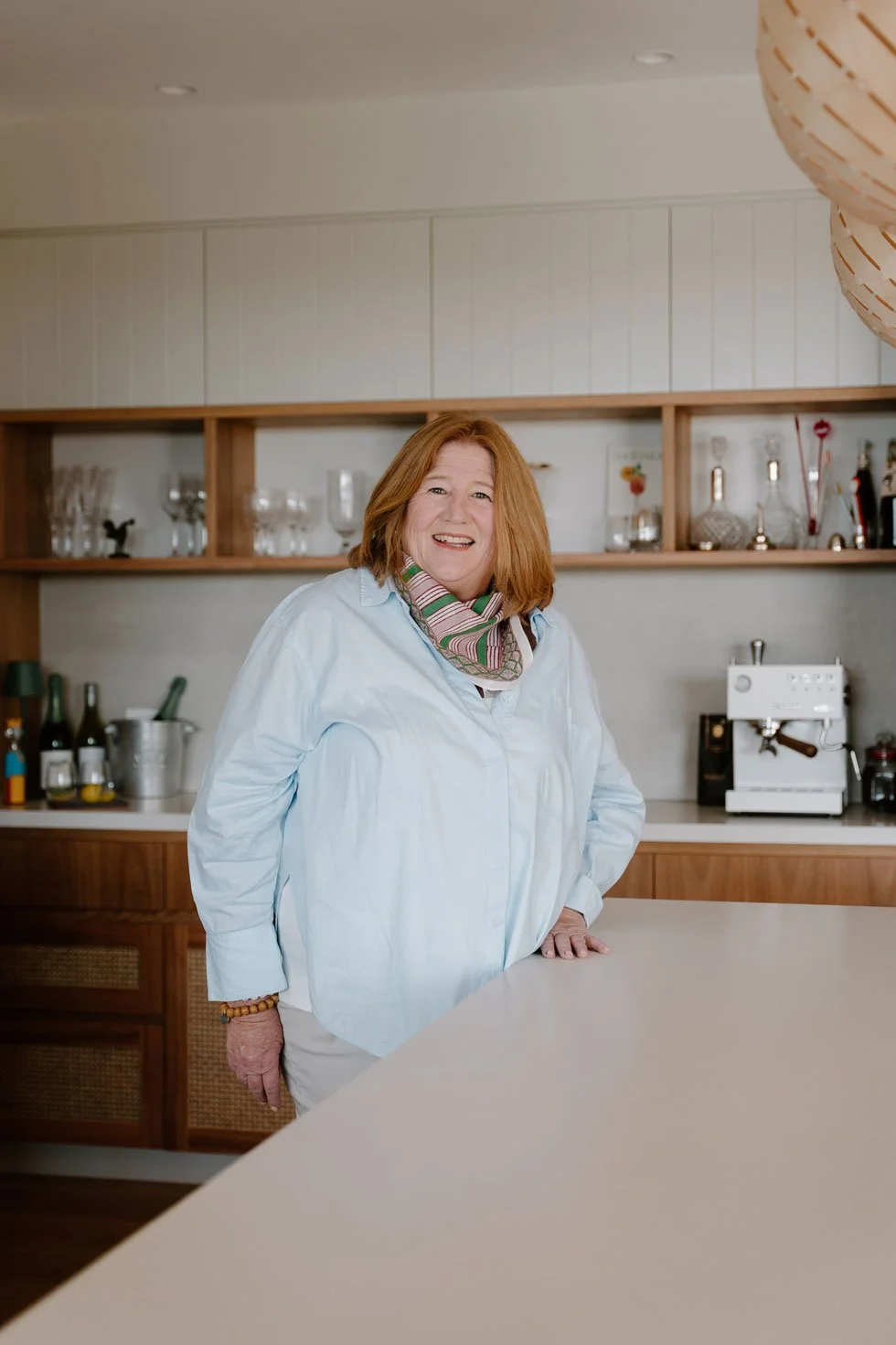 Suzie, smiling, wearing a colourful scarf standing in a modern kitchen, resting her hand on a white countertop.