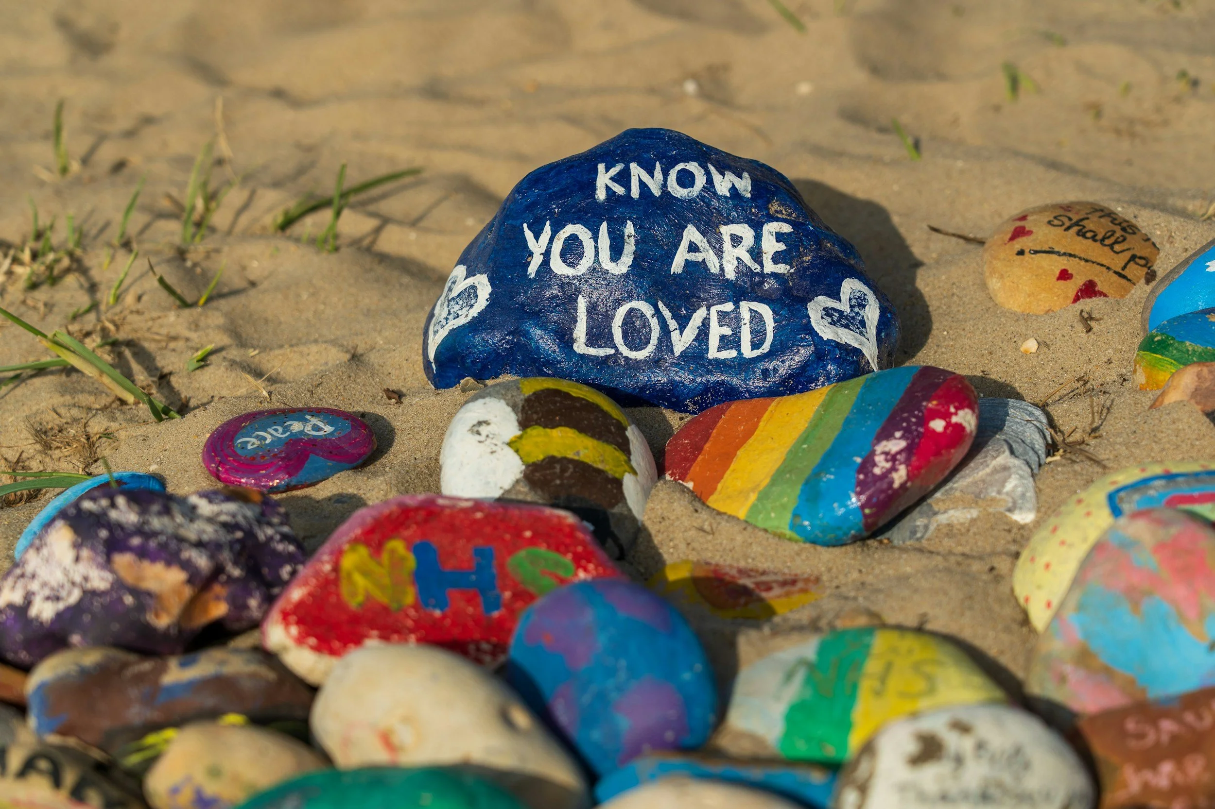 Colorfully painted rocks on sand, with one large blue rock inscribed 'KNOW YOU ARE LOVED' and various other decorated rocks with messages and rainbow patterns.