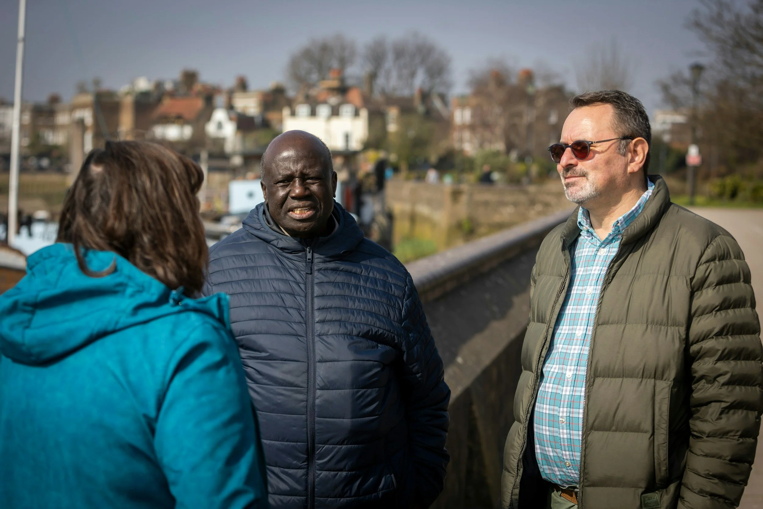 Three adults engaged in conversation outdoors on a sunny day, with a row of houses and trees in the background.