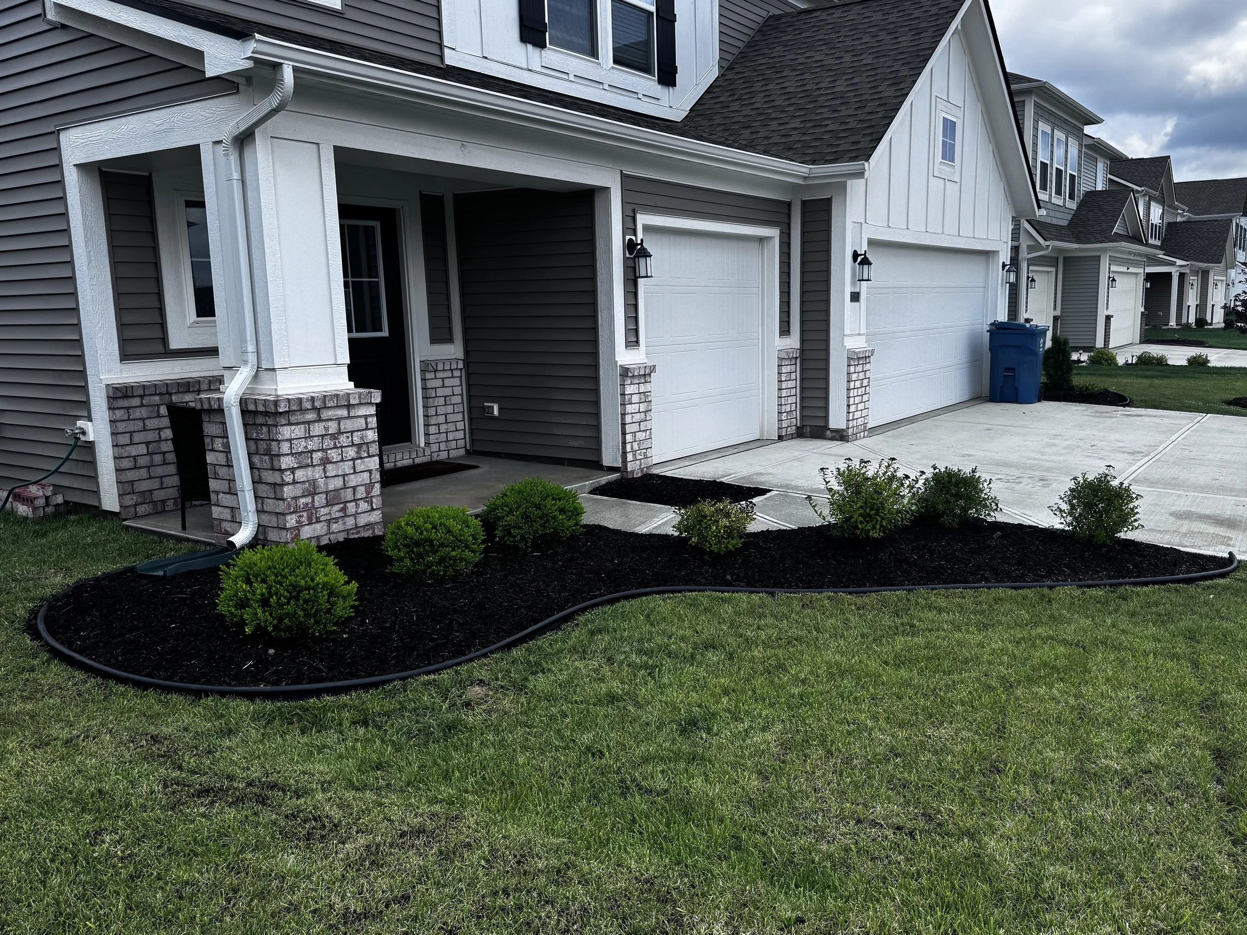 Modern residential house with a manicured front yard, black mulched flower bed with small green shrubs, concrete driveway, attached garage, gray siding, white trim, and a blue trash bin.