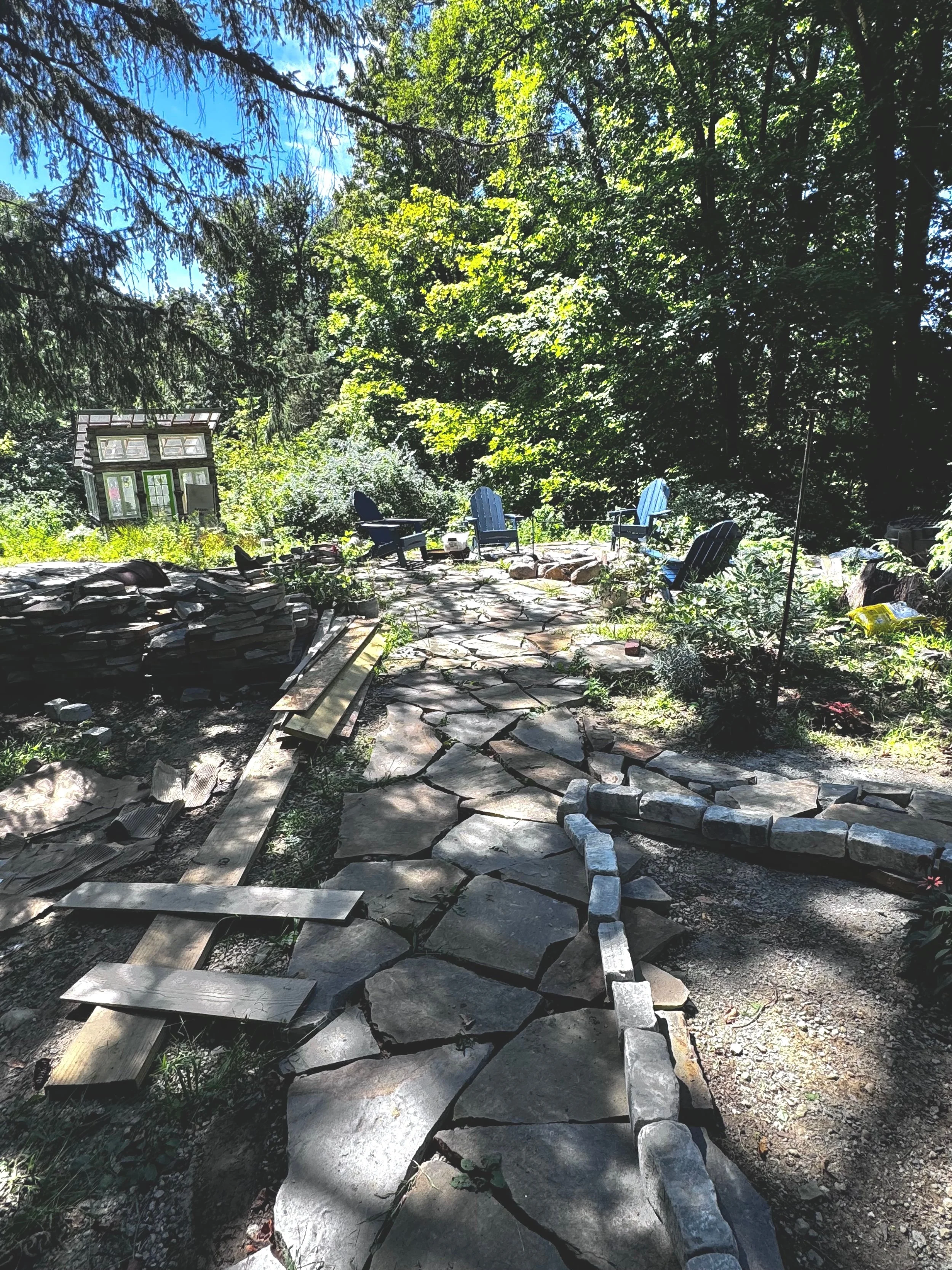 A backyard with a stone pathway under construction, surrounded by trees and outdoor seating, with blue Adirondack chairs and a small structure in the background.