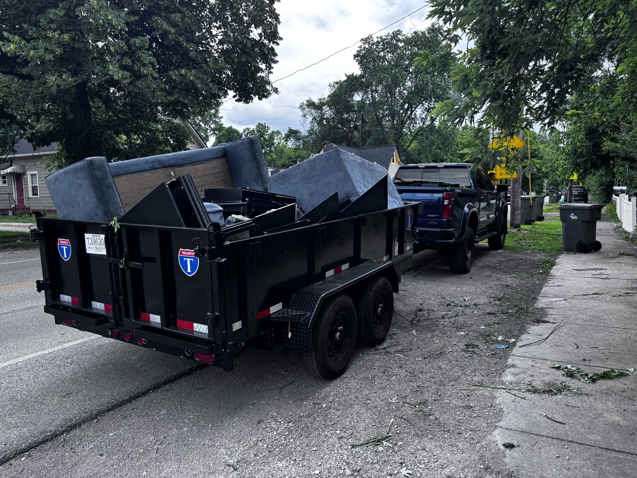 A truck with a trailer carrying a displaced couch and other furniture on a residential street with trees, houses, trash cans, and street signs in the background.