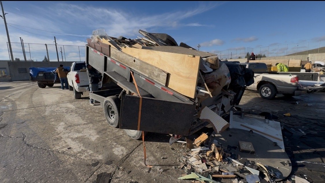 A garage filled with a trailer loaded with furniture and household items, with an open garage door revealing a driveway and suburban houses outside.
