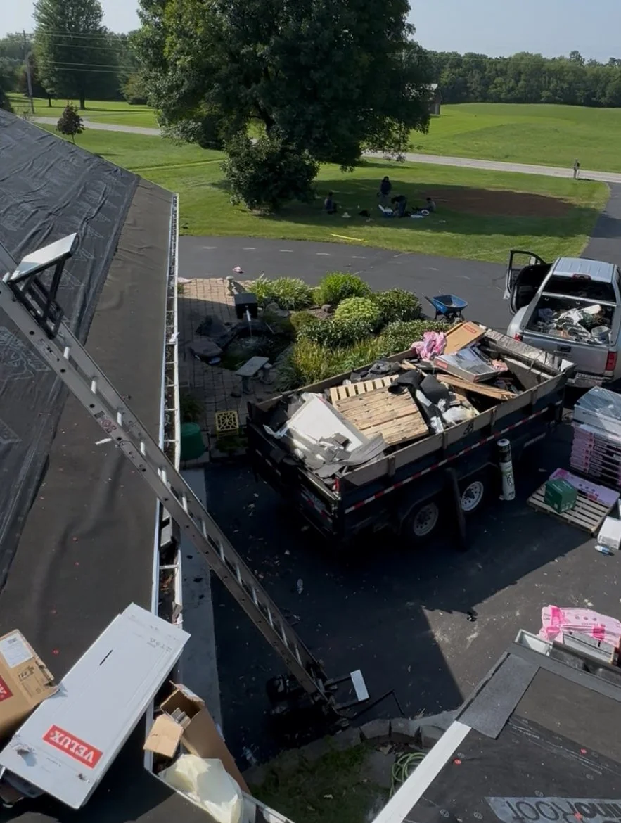 A garage filled with a trailer loaded with furniture and household items, with an open garage door revealing a driveway and suburban houses outside.