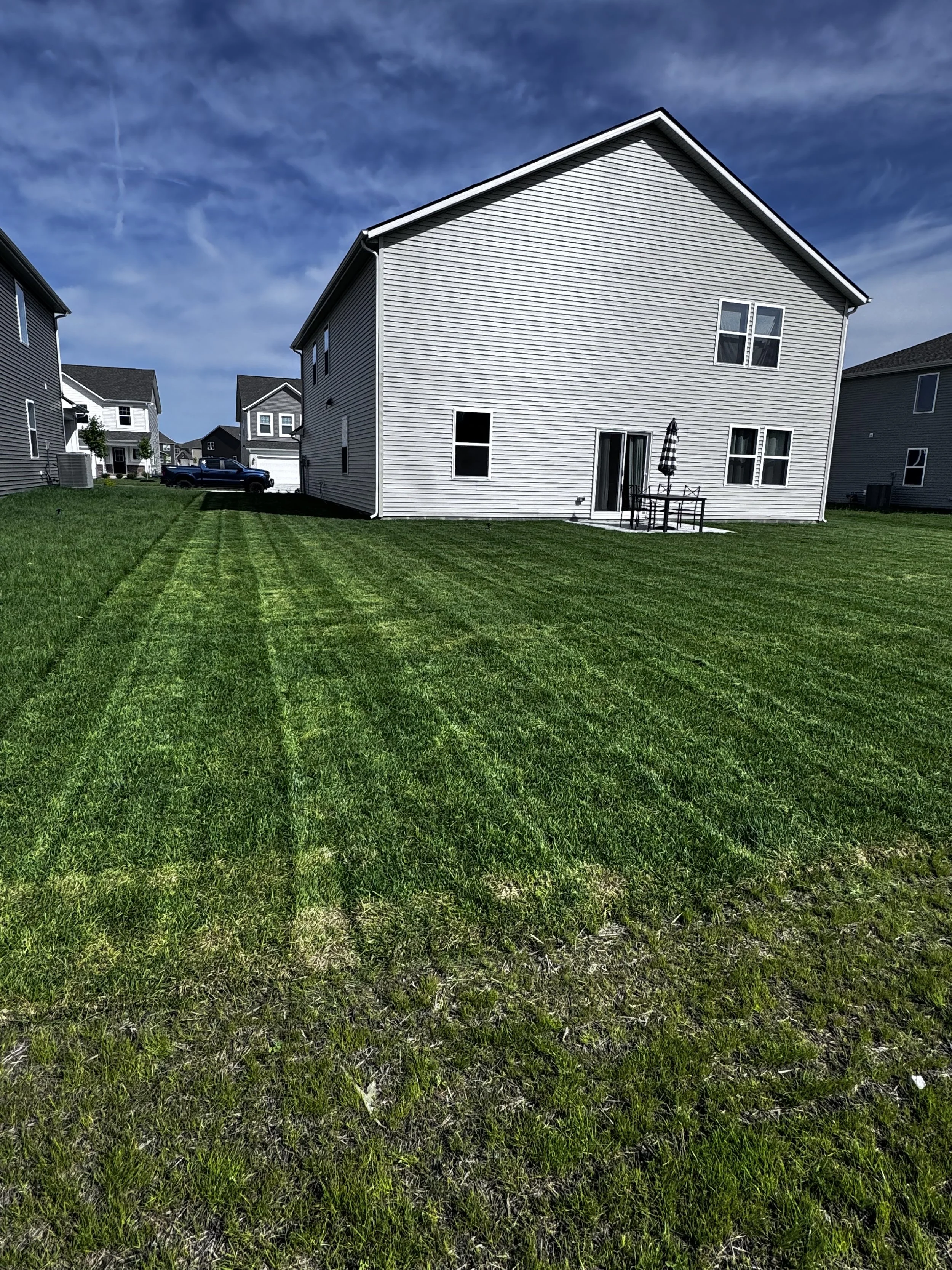 Backyard with green grass, house with white vinyl siding, multiple windows, patio table with umbrella, and neighboring houses visible under a blue sky with some clouds.