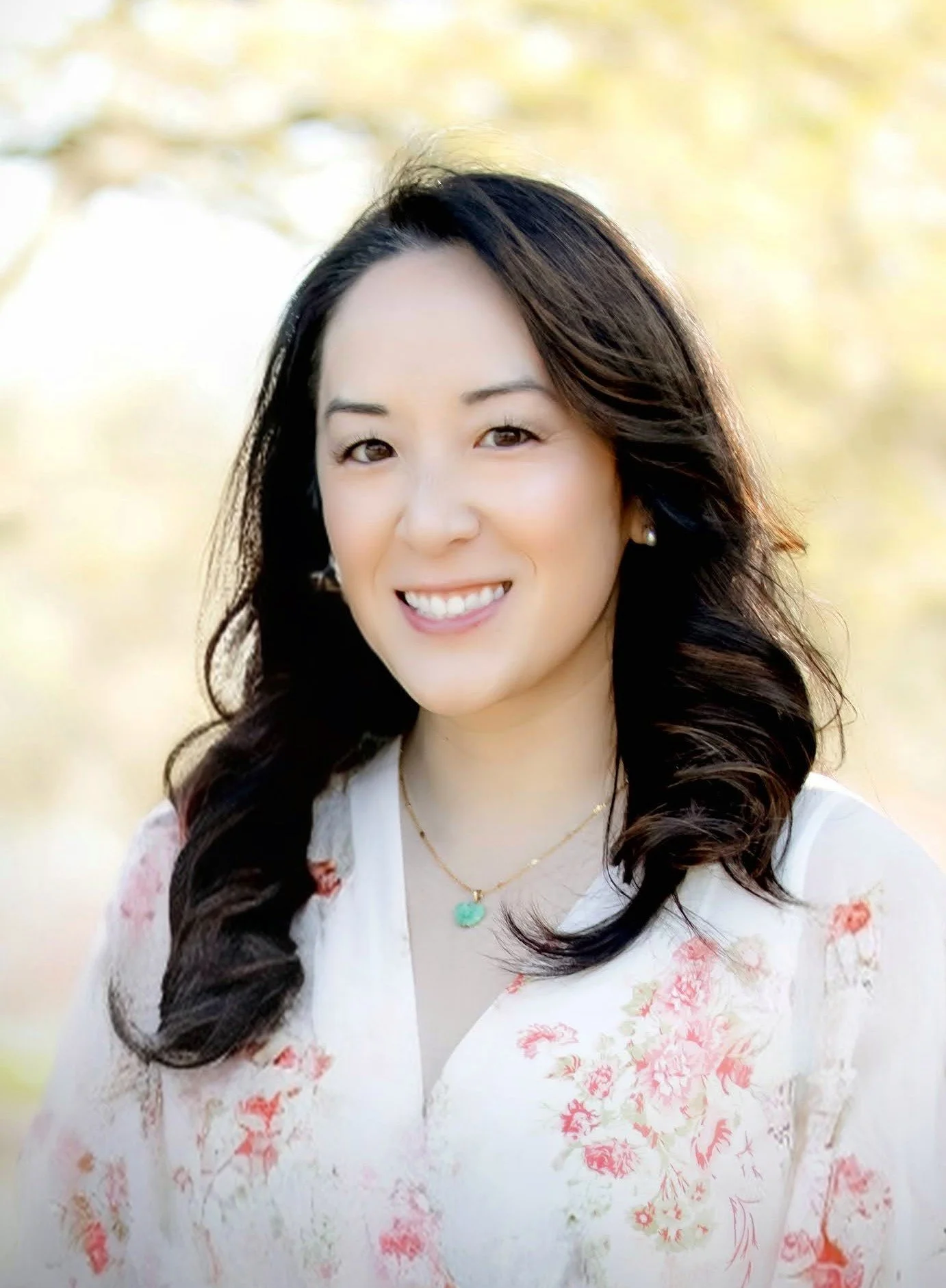 A smiling woman with dark wavy hair wearing a white floral top, a gold necklace with a turquoise pendant, and pearl earrings, outdoors with blurred trees in the background.