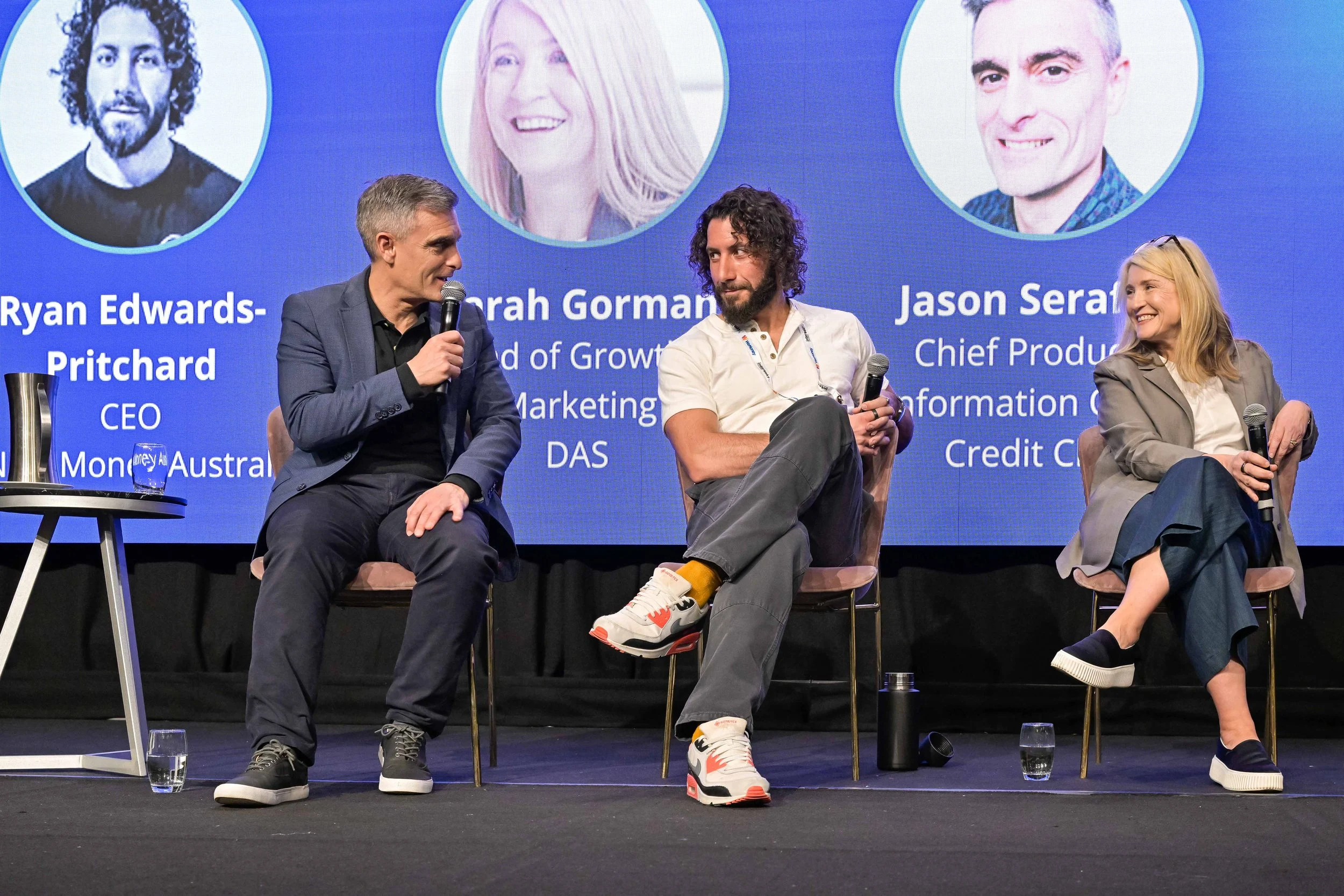 Three people sitting on stage participating in a panel discussion, with a blue background displaying their names and titles.