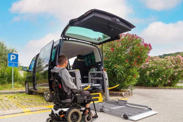 A person in a wheelchair using a wheelchair lift to enter a black van in a parking lot near pink flowers and greenery.
