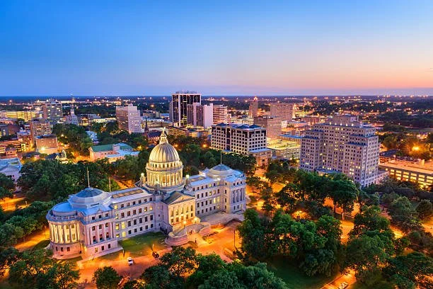Aerial view of downtown Montgomery, Alabama at sunset, with the Alabama State Capitol building in the foreground and city lights illuminating surrounding buildings.