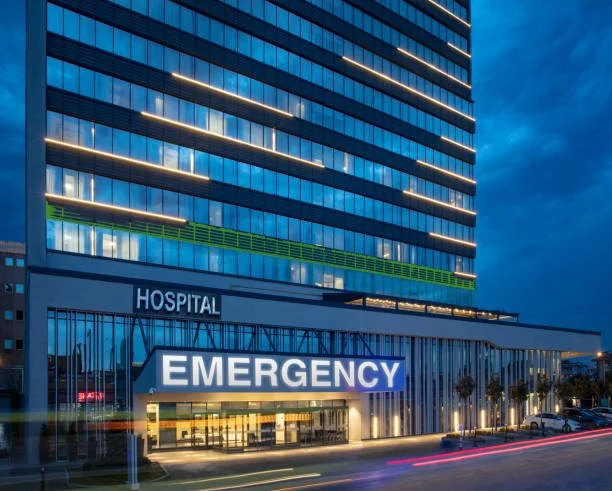 Modern hospital emergency entrance with a tall glass building above, illuminated signs reading 'HOSPITAL' and 'EMERGENCY', at dusk with light trails from passing cars.