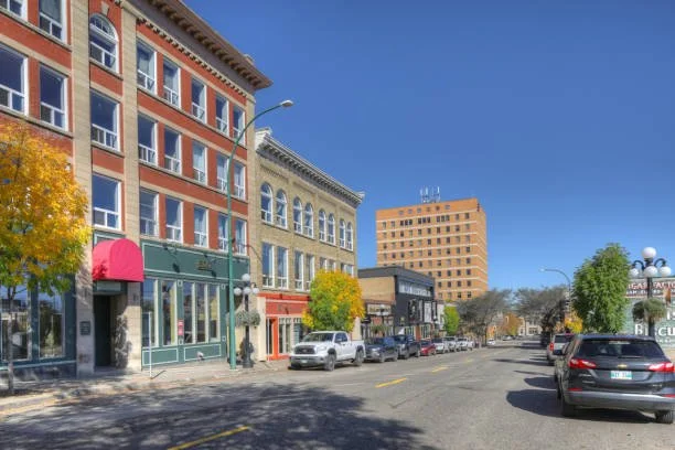 Downtown street with multistory buildings, parked cars, and trees with fall foliage under a clear blue sky.