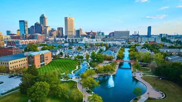 A city skyline with tall buildings, a park with a pond, walking paths, and green trees in the foreground on a clear day.