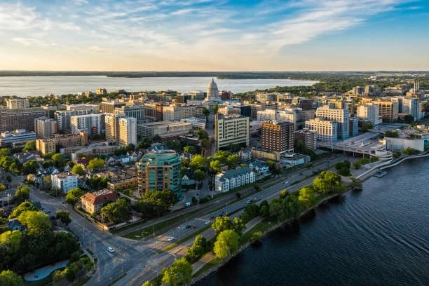 Aerial view of a cityscape featuring office buildings, residential areas, a river in the foreground, and governmental buildings, with a bridge on the right and a large body of water in the background during sunset.
