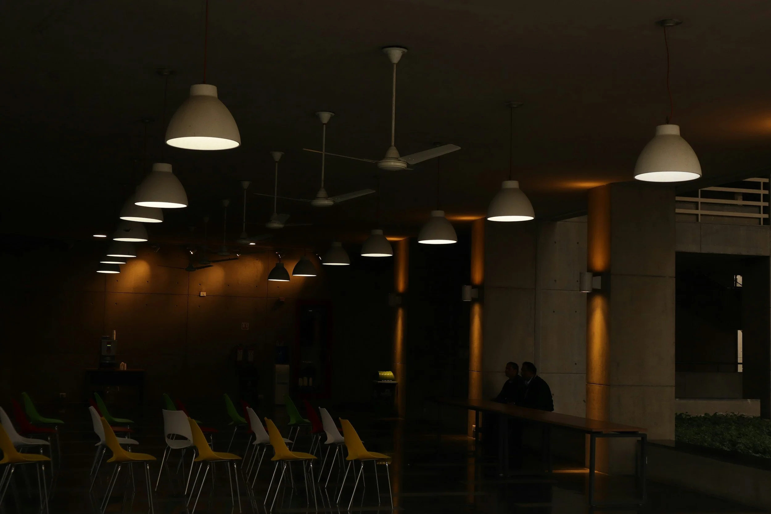 A dimly lit modern indoor space with white pendant lights hanging from the ceiling, ceiling fans, and colorful chairs arranged around tables, with a warm glow on the walls and columns.