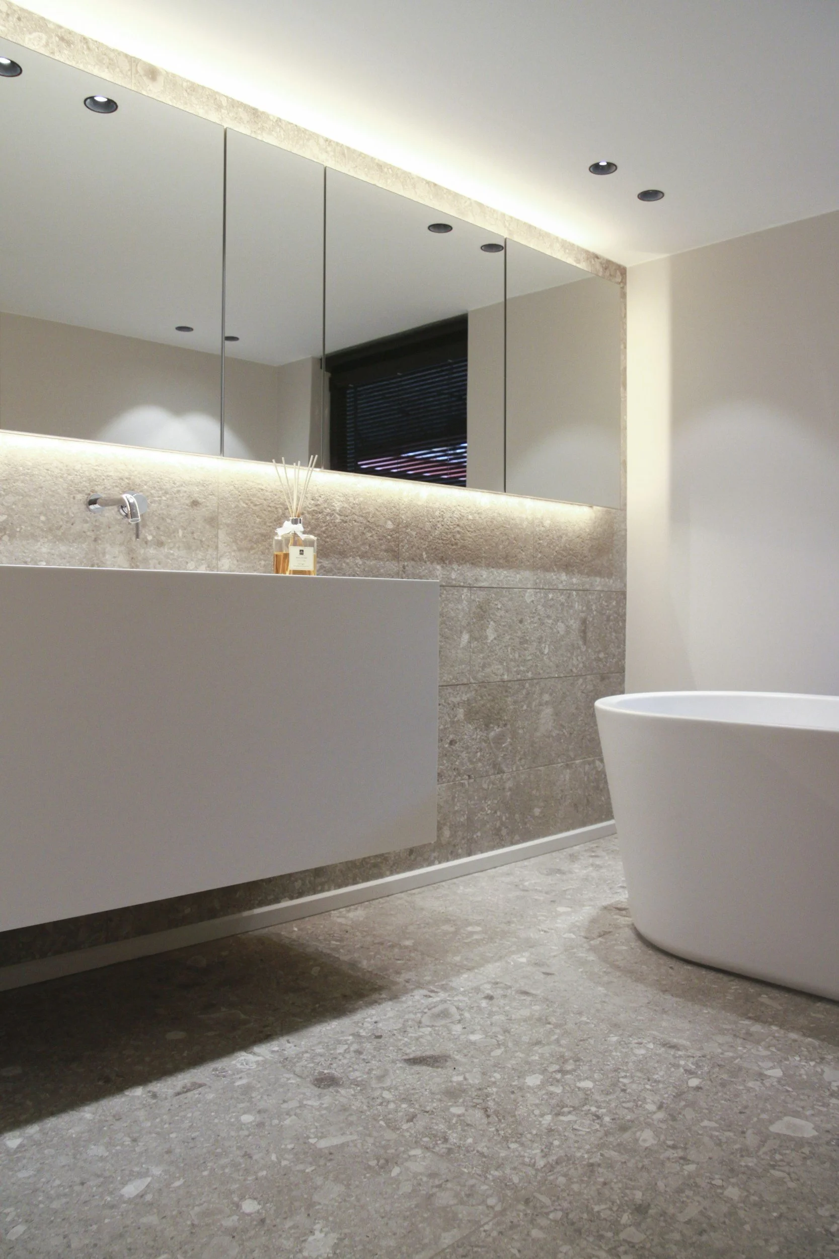 Modern bathroom with a large mirror, beige textured walls, a white freestanding bathtub, and a floating vanity with a bottle of fragrance on top. Recessed ceiling lights and a window with blinds are visible.