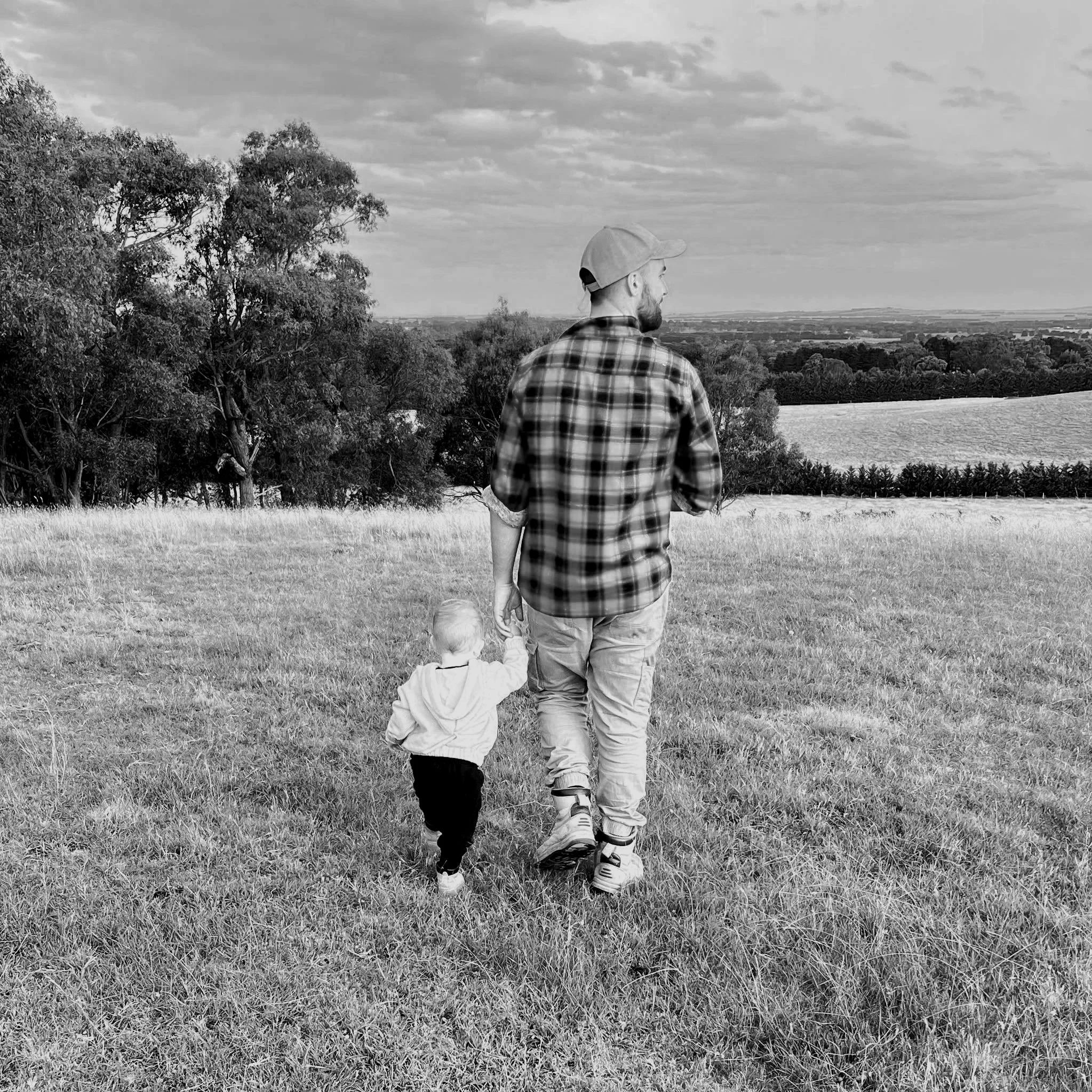 A man and a child walking through a field, with trees and a landscape in the background under a cloudy sky, in black and white.