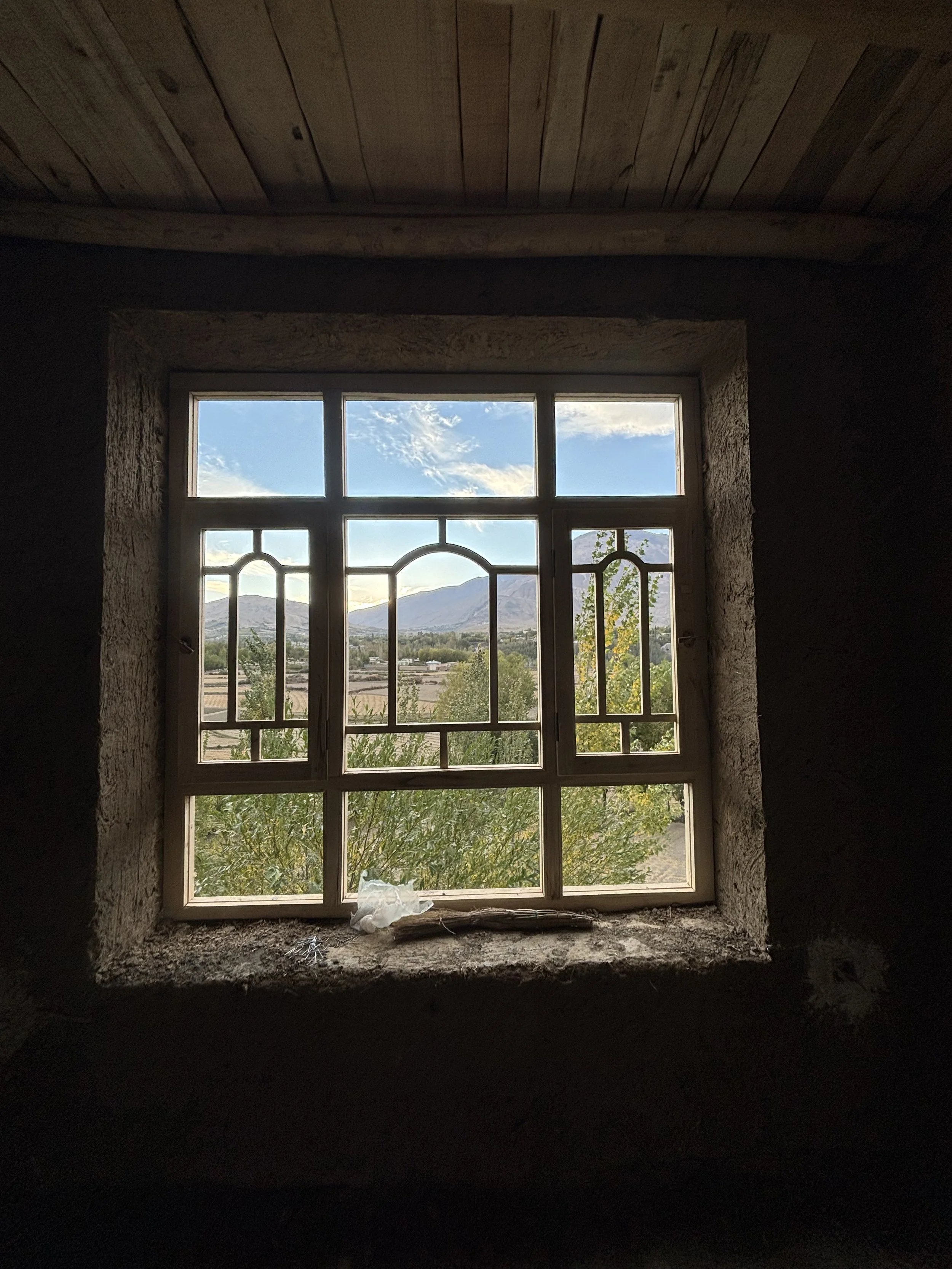 View of a window with natural scenery outside, including mountains, trees, and blue sky with clouds, framed by rough stone wall and ceiling inside an Afghan home.