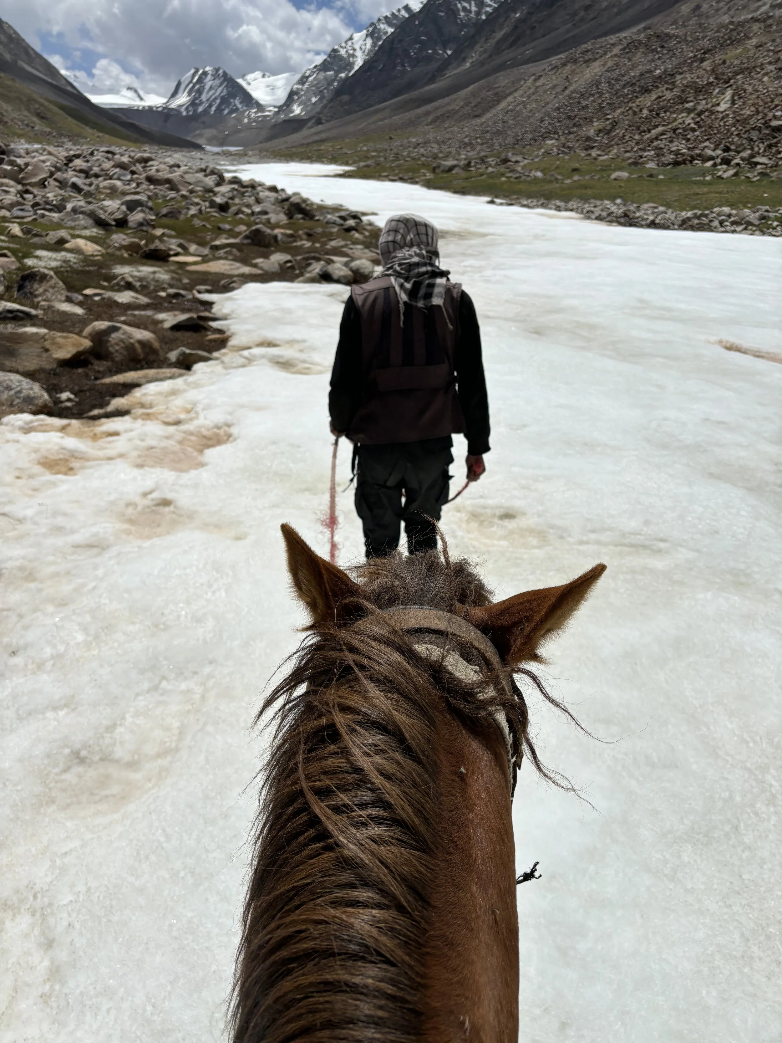 A person walking along a snowy river in a mountainous landscape, seen from behind on horseback, with snow-capped peaks in the distance in Afghanistan.