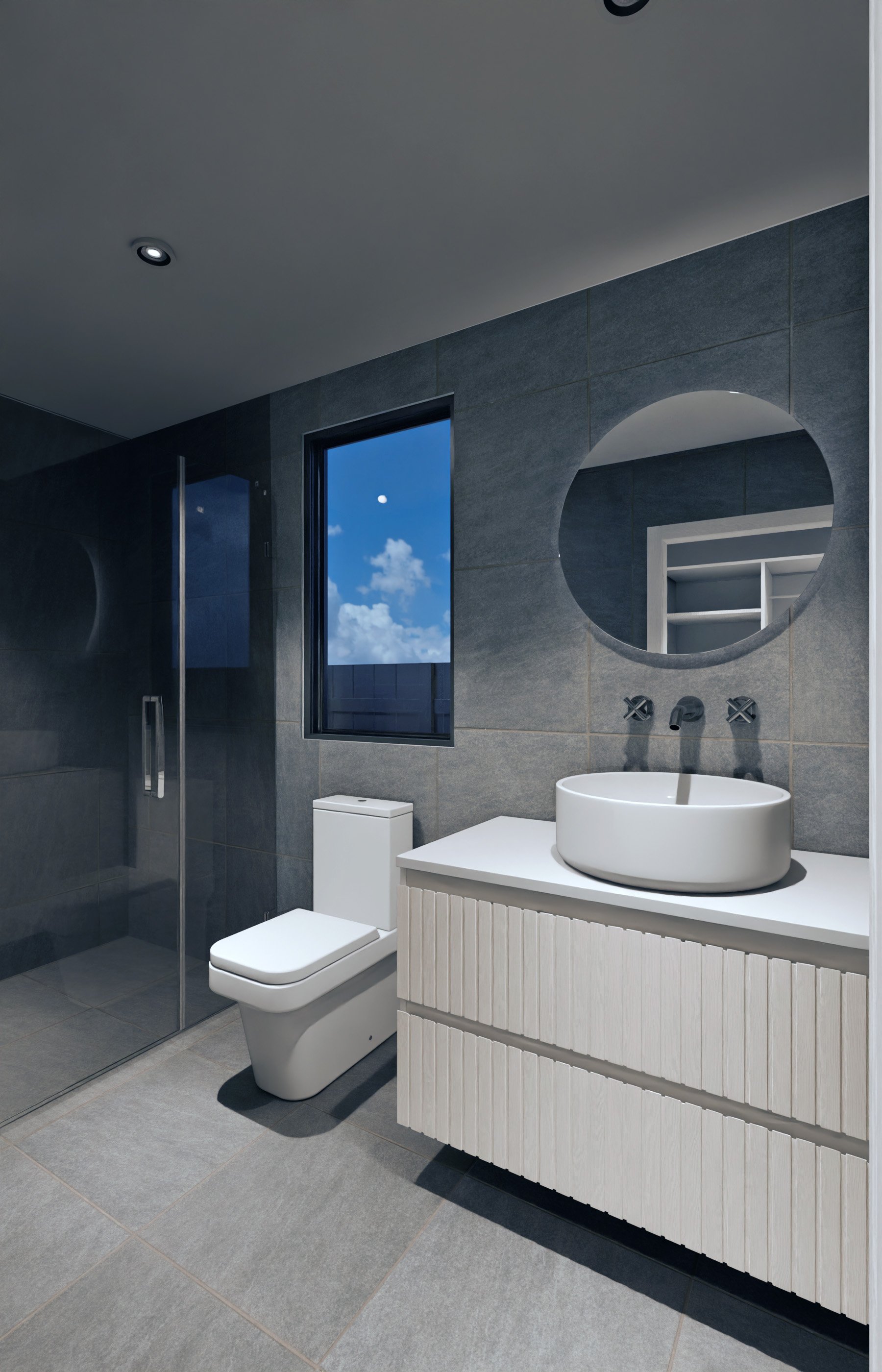 Modern bathroom with gray tiled walls and floor, a white toilet, a white vanity with a round vessel sink, a large round mirror, a window showing a blue sky with clouds, and a glass-enclosed shower.