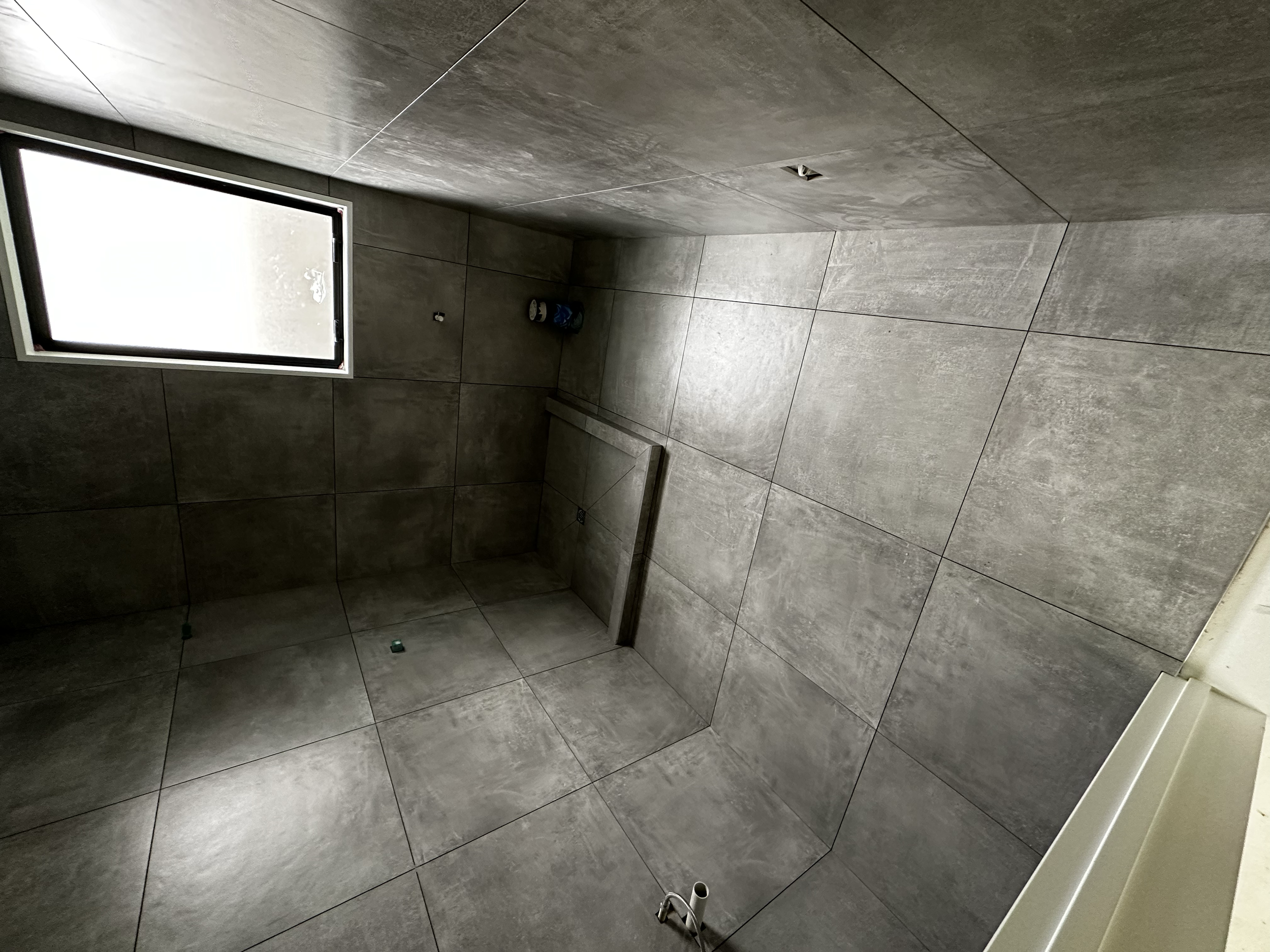 Empty bathroom with large gray tiles on walls and floor, a small window, and plumbing fixtures for a toilet and sink not yet installed.