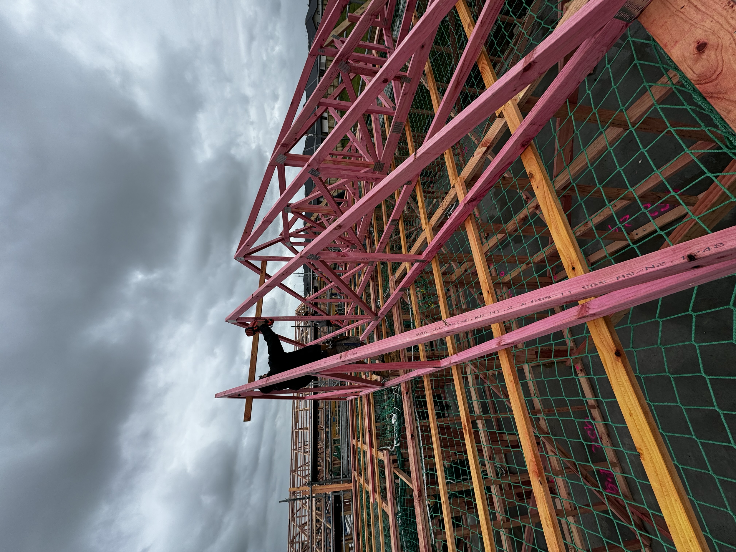 Construction worker installing pink wooden trusses on a building under construction with cloudy sky in the background.