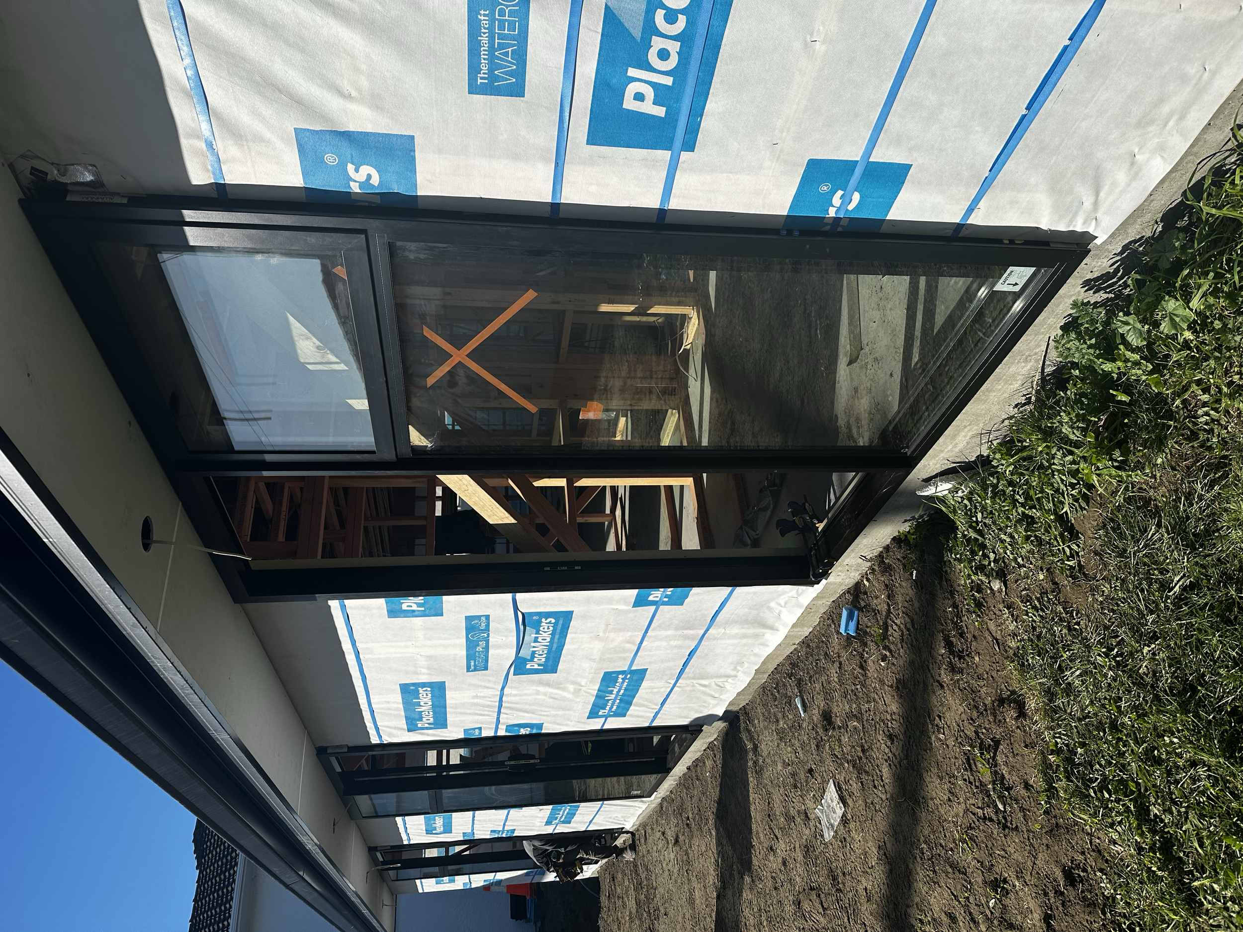Partially constructed screened porch or sunroom with black framing and large glass panels, adjacent to house wrapped in water-resistant building wrap, next to green shrubbery and soil.