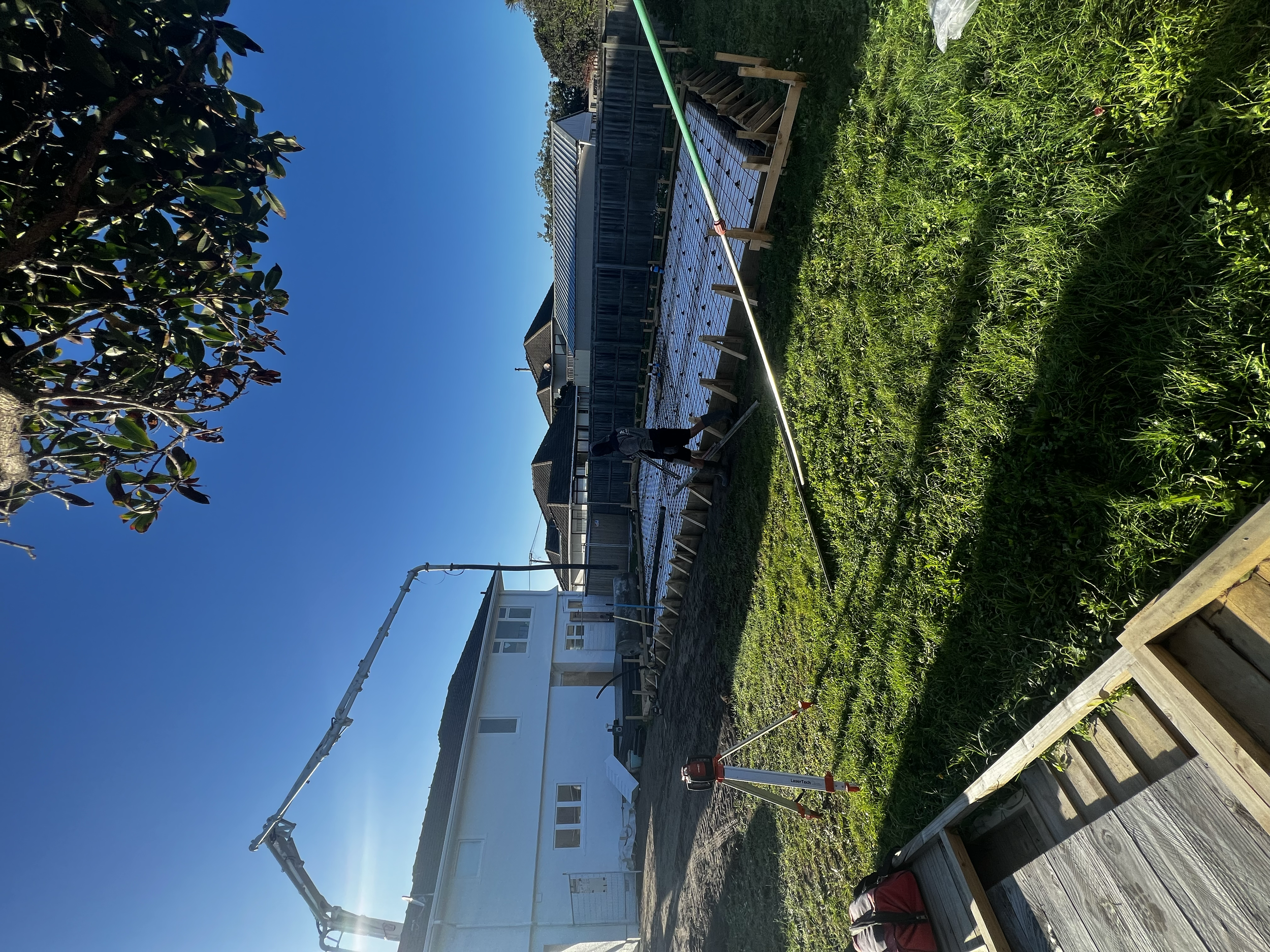 Construction workers are laying concrete in a backyard with a white house, green grass, and a blue sky. A concrete pump truck is pouring concrete, and one worker is guiding the flow while another is nearby.