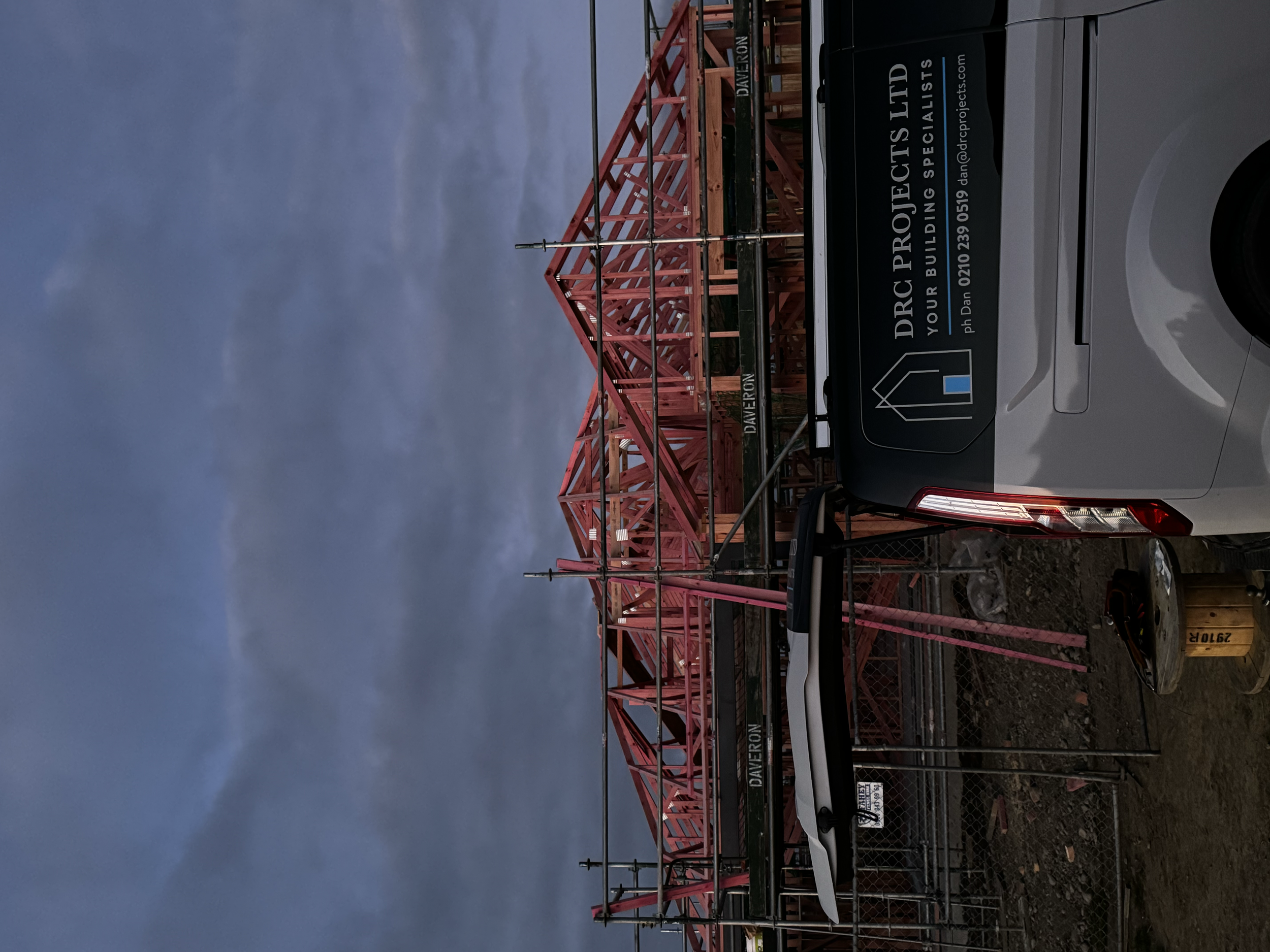 Construction site with a red steel framework and a white van with an advertisement for DRC Projects Ltd, under a cloudy sky.