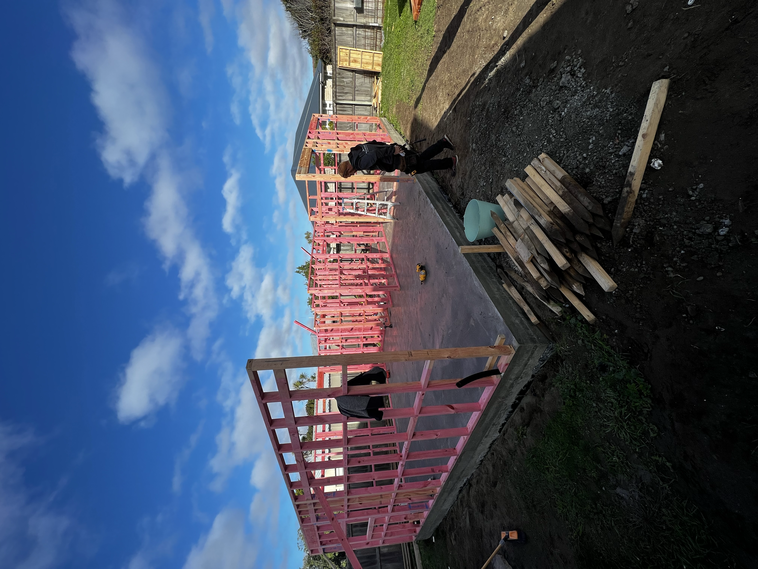 Construction site with a wooden frame of a house painted pink, and two people working on the build, under a blue sky with clouds.