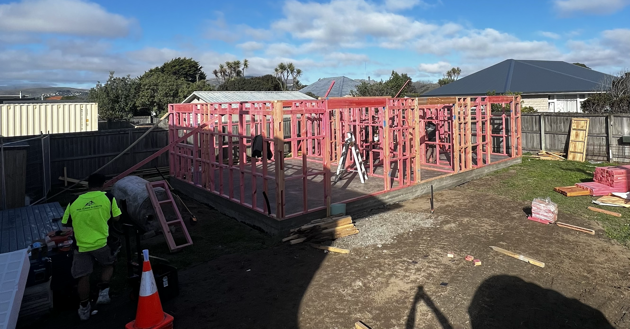 Construction site with a wooden framing structure for a house, workers present, pink insulation panels stacked on the ground, and neighboring houses in the background.