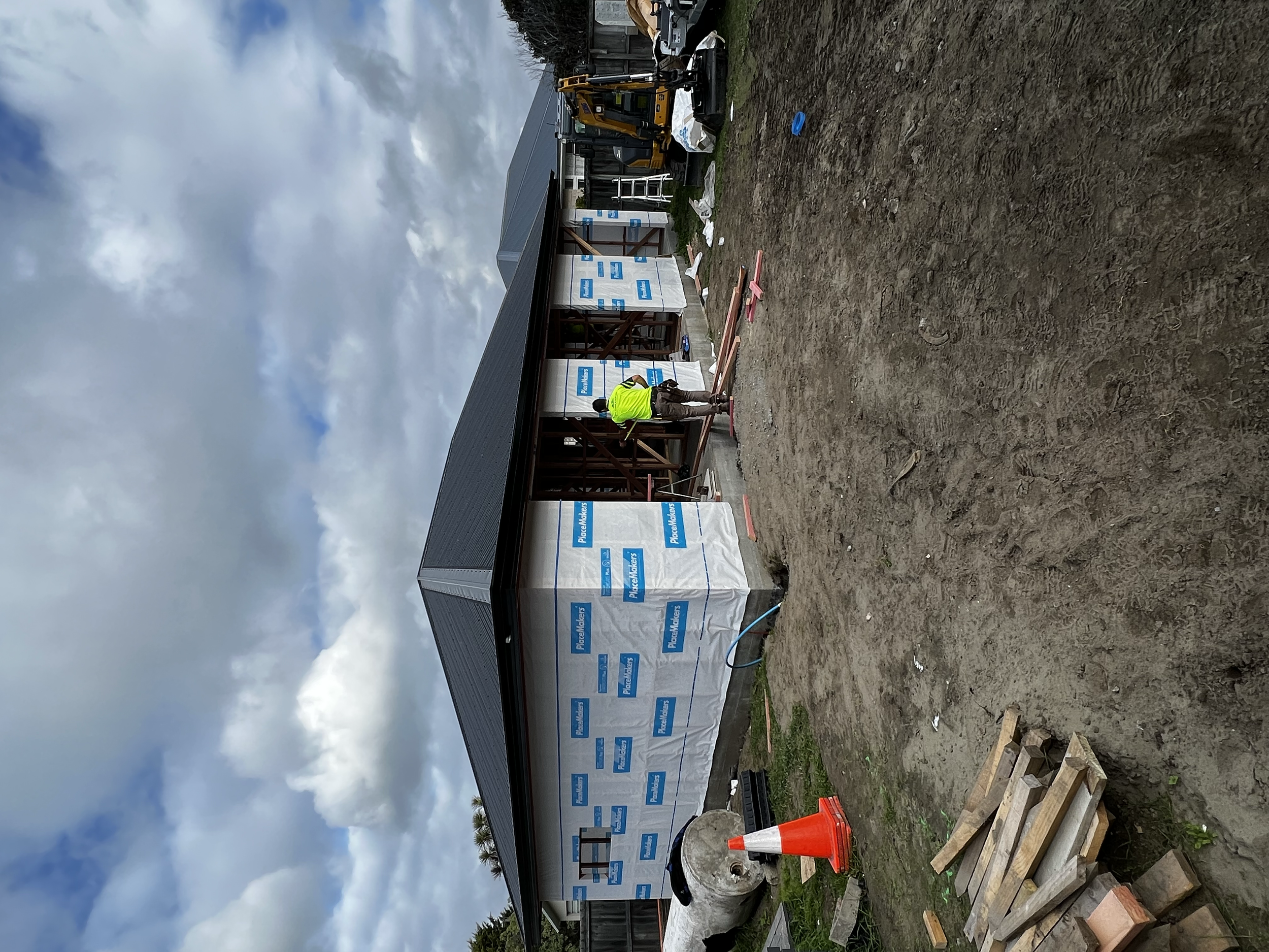 Construction worker installing siding on a house under a cloudy sky, with building materials and equipment on a dirt ground.