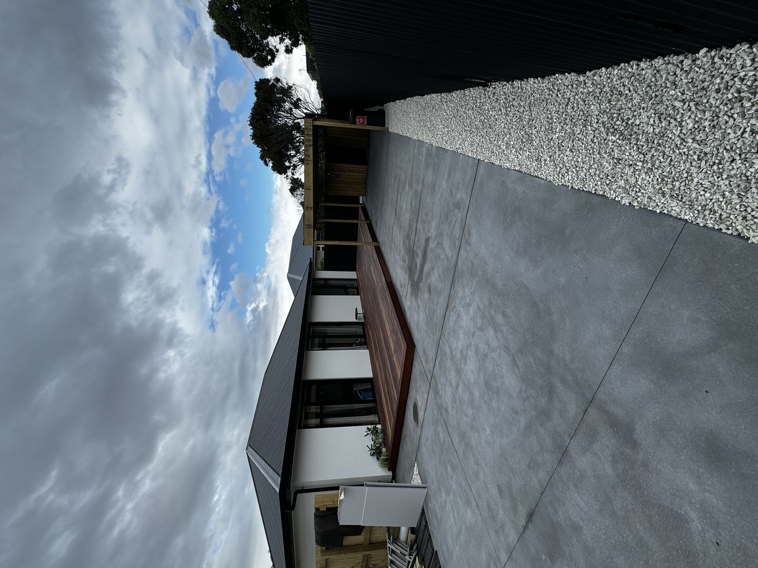 Modern house with grey roof, white walls, and a small deck, surrounded by a gravel and concrete yard, under cloudy sky.
