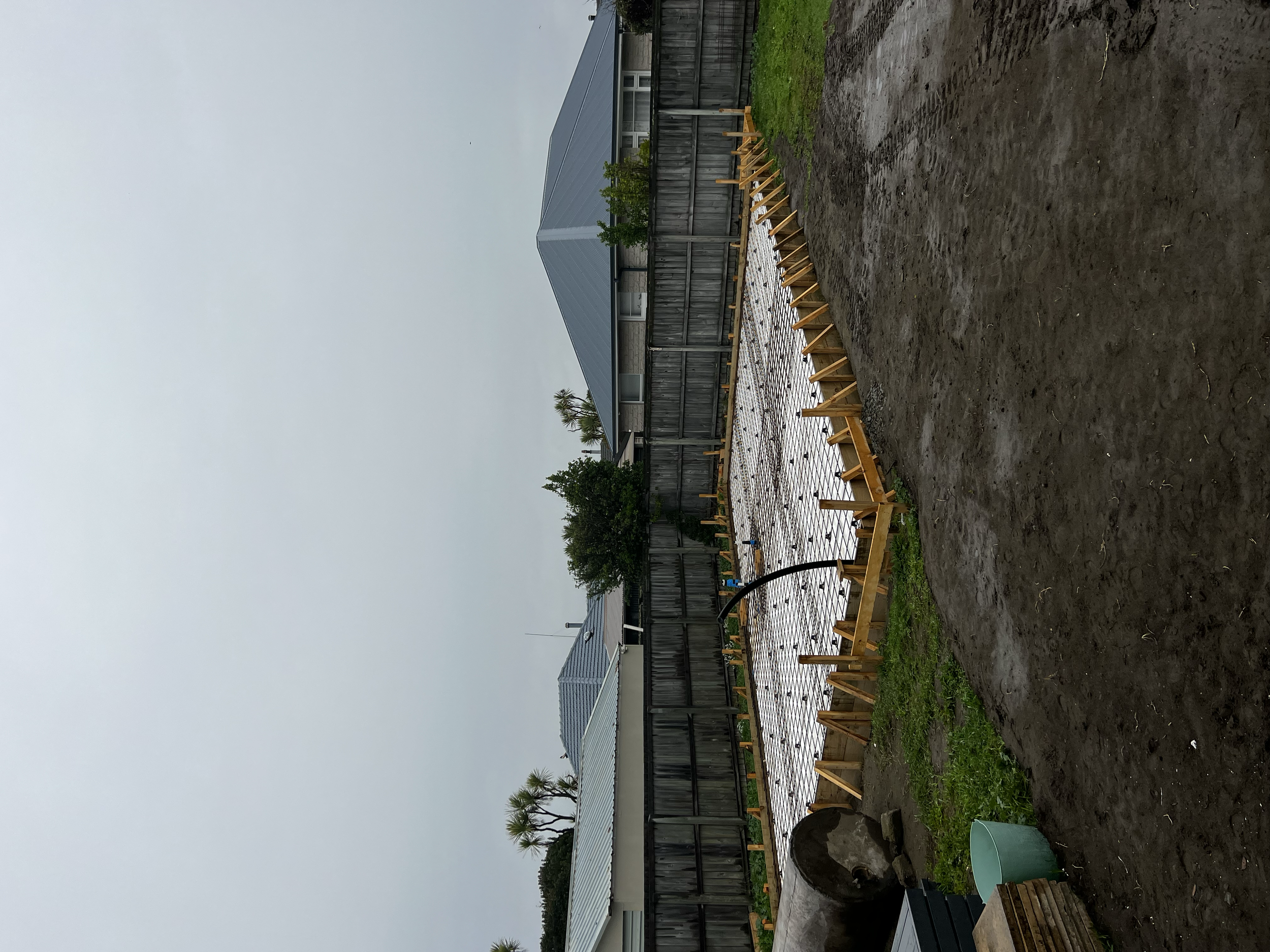 Backyard with garden bed under construction, surrounded by a wooden frame, with a house in the background, cloudy sky, and some trees.