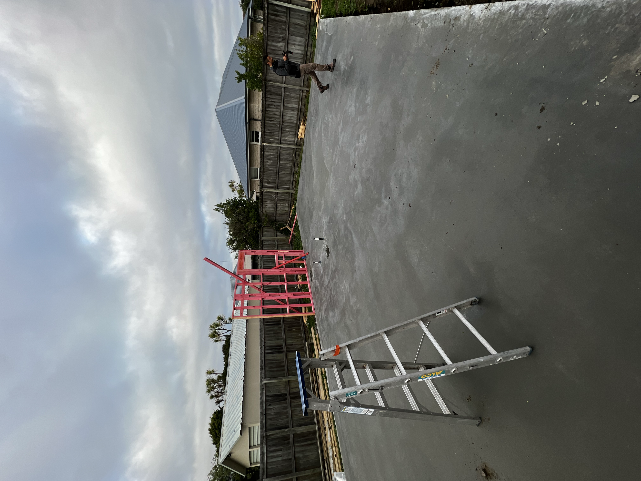 Construction site with a concrete floor, a pink partially built structure, a silver ladder, and a man walking in the background.