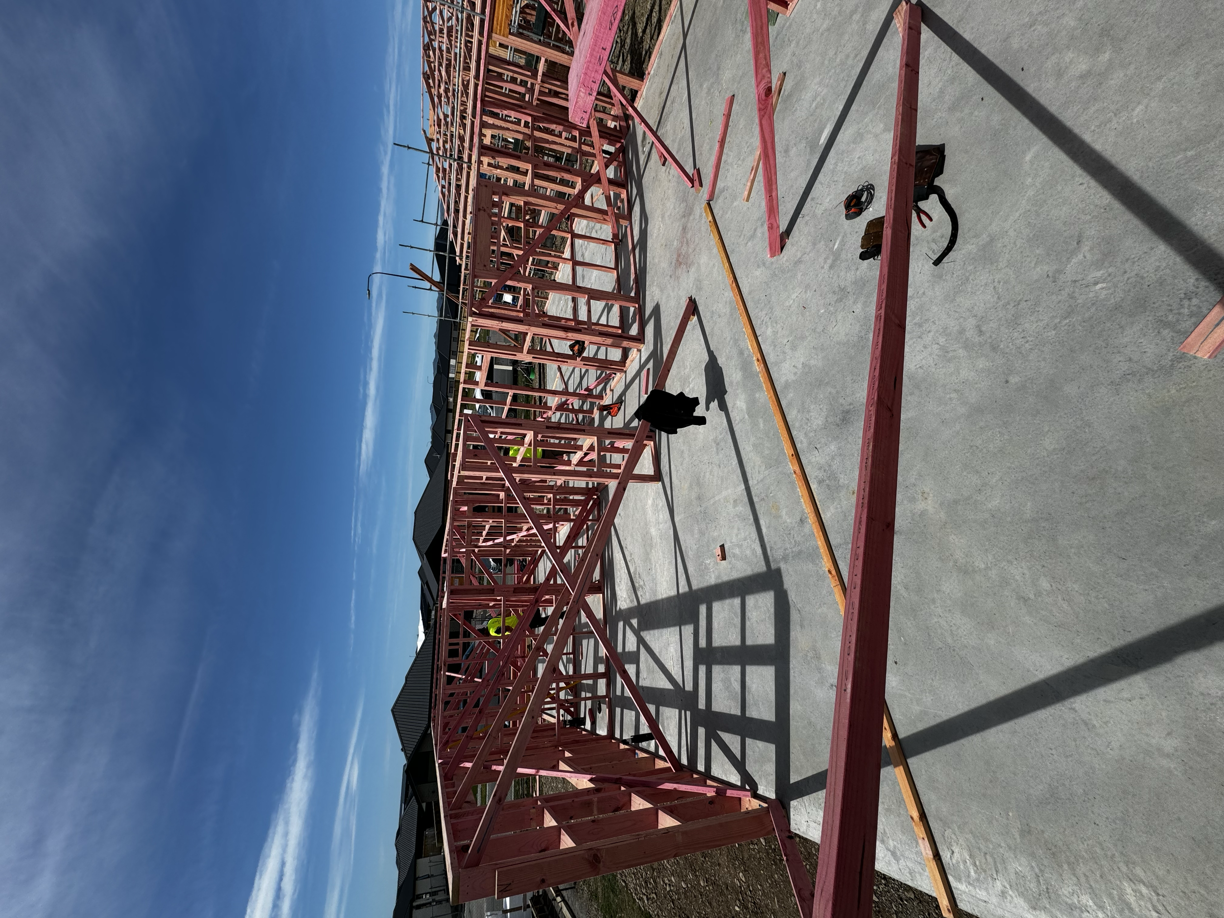 Construction site with a wooden framing structure on a concrete foundation, blue sky with some clouds, and construction tools on the ground.