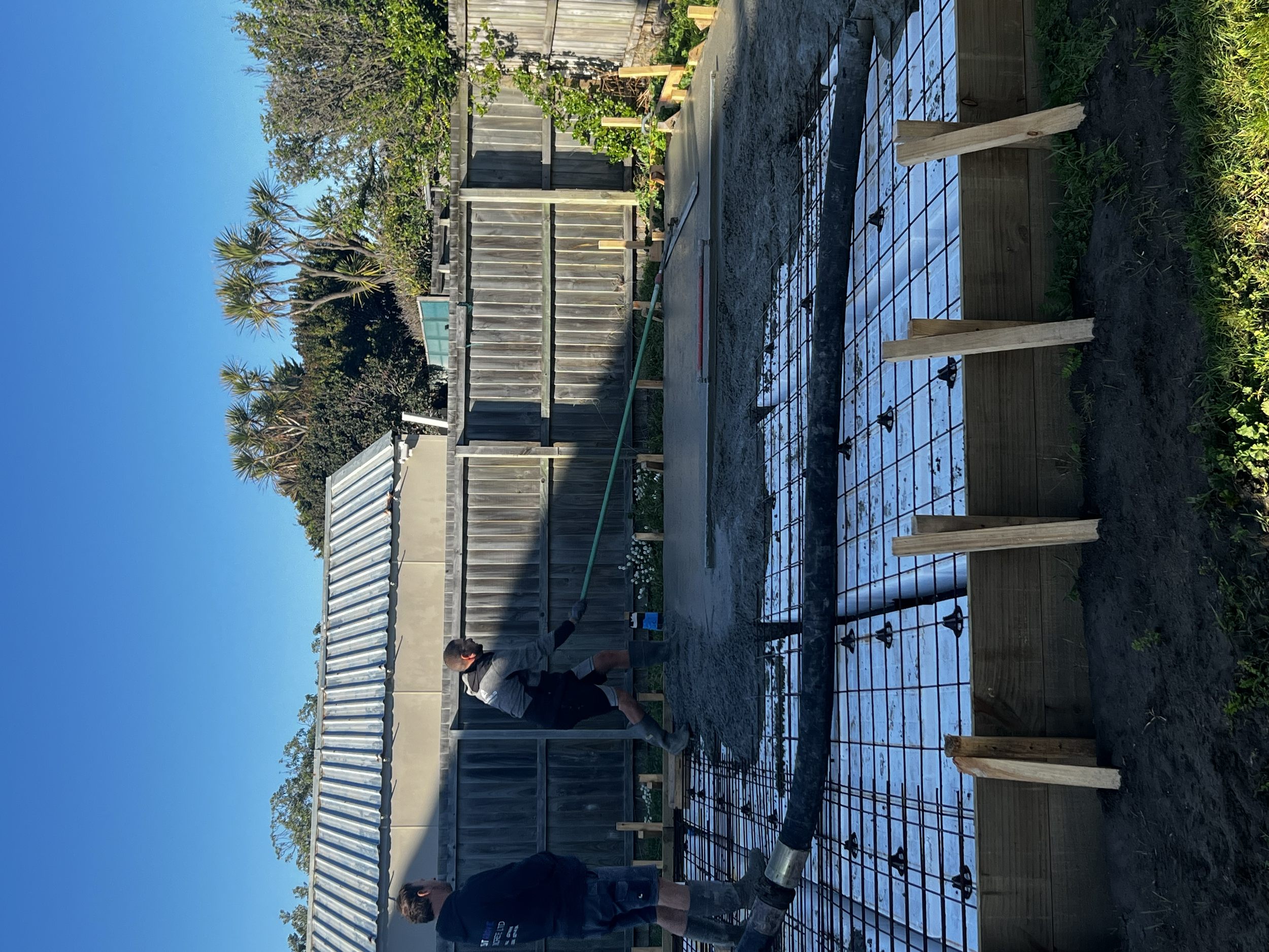 People working on installing a foundation for a building or structure, with rebar grid and wooden supports, in a backyard with fencing and trees.