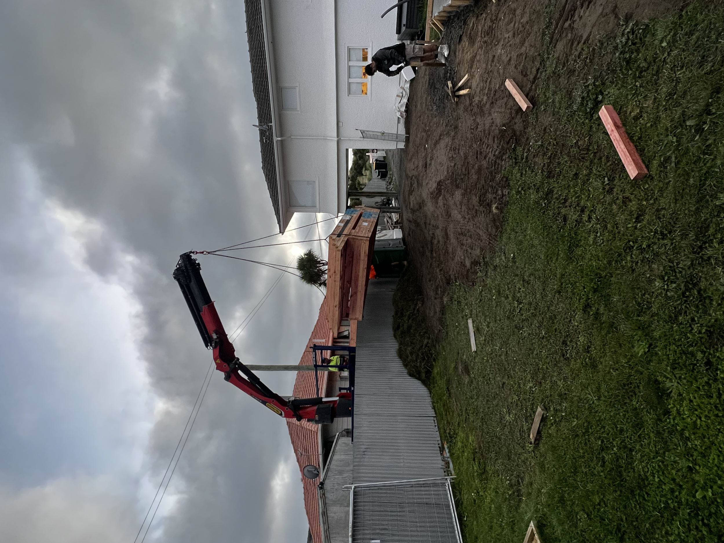 A construction worker operating a crane on a residential house construction site under cloudy skies, with a grassy area and building materials in the foreground.