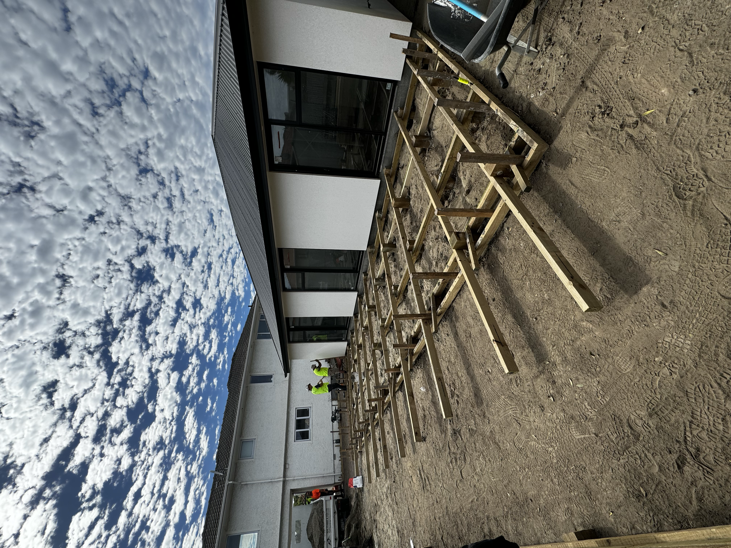 Construction site of a residential building with wooden framing and two workers in safety vests. The building has large glass windows and a black roof. The sky is partly cloudy.