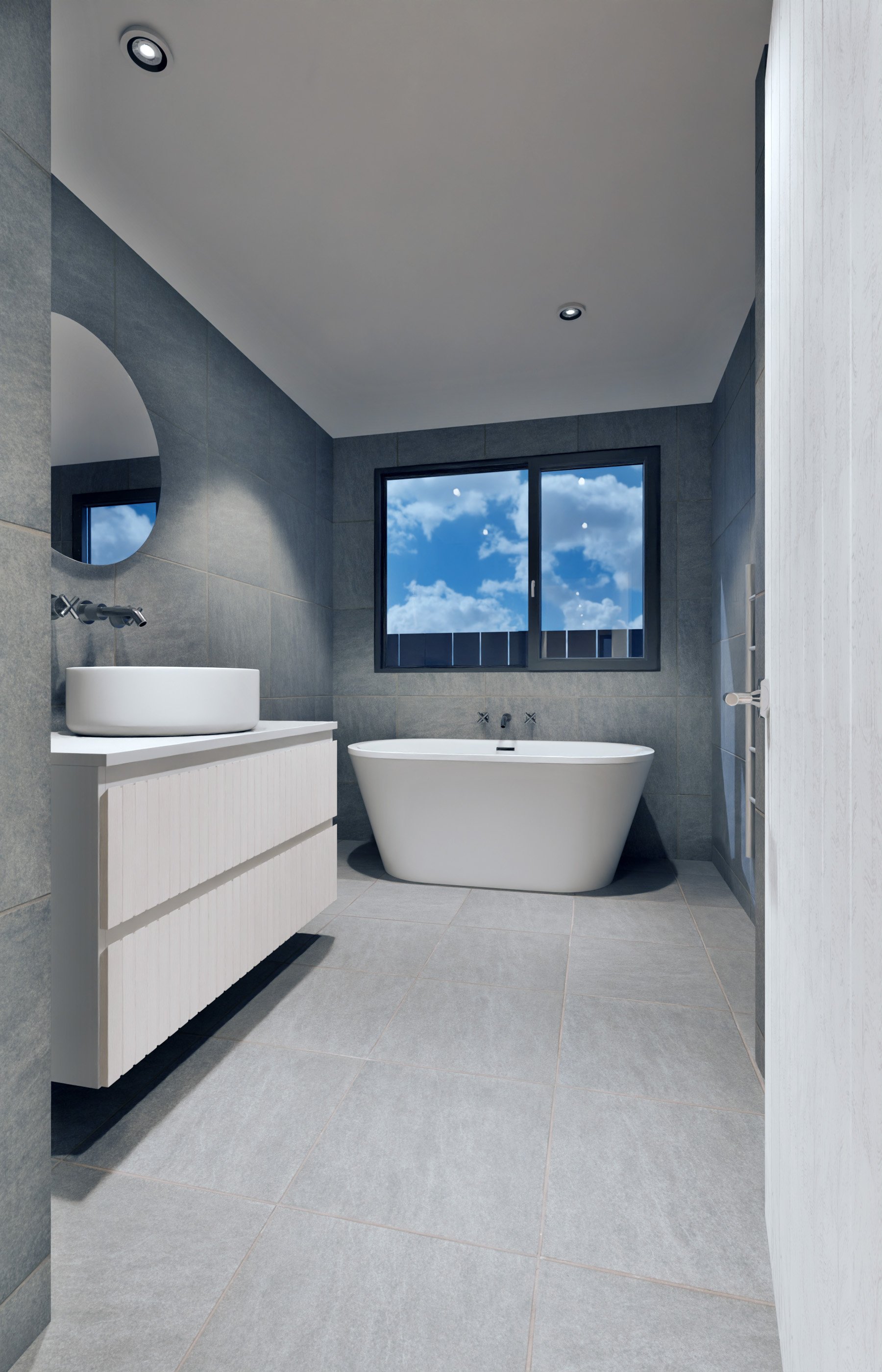 Modern bathroom with gray tiled walls and floor, a white freestanding bathtub under a window showing a blue sky with clouds, a white vanity with a vessel sink, a round mirror, and a towel radiator.