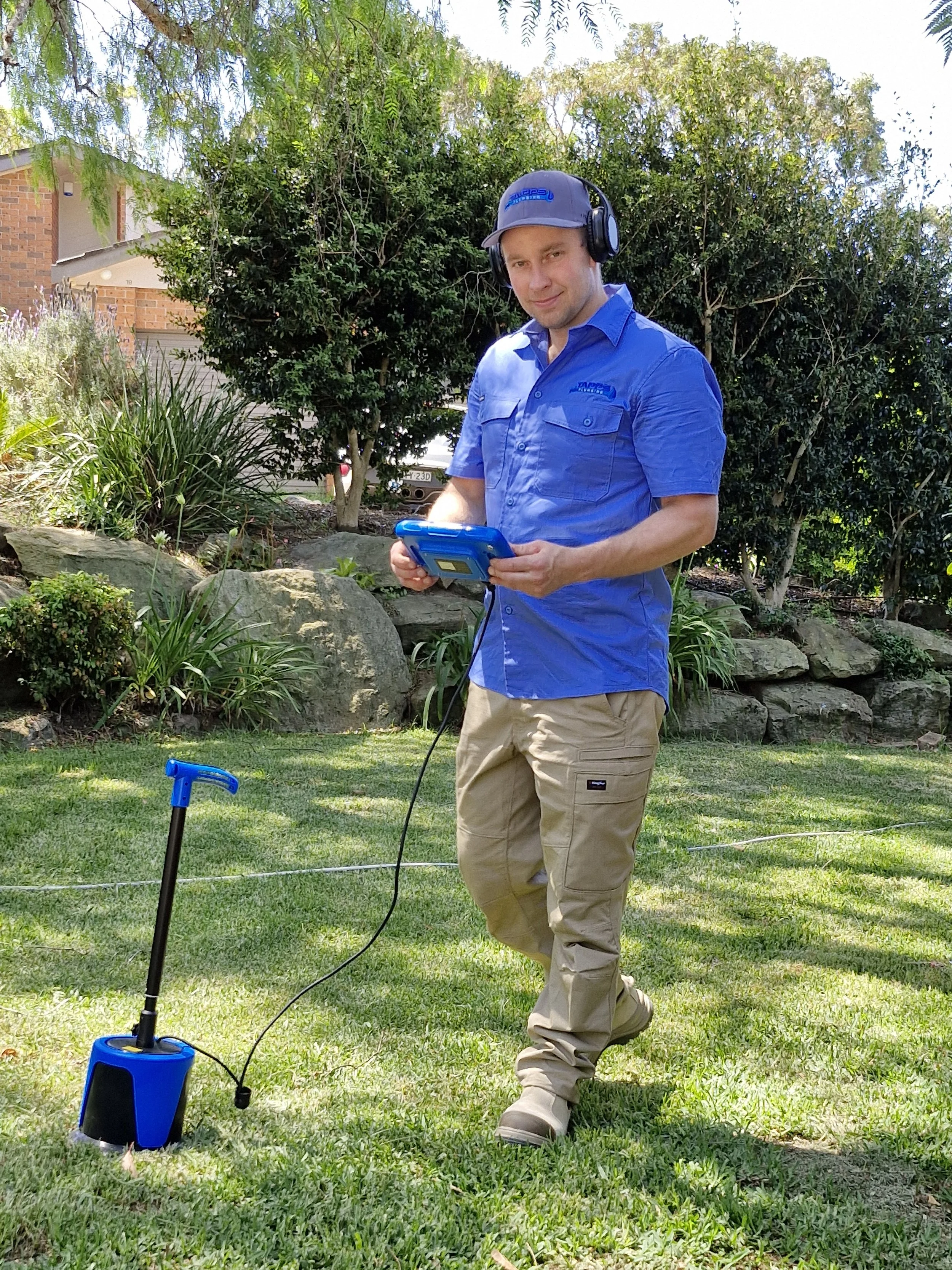 A man in a blue shirt and khaki pants measuring the ground with a metal detector in a garden with trees and bushes.