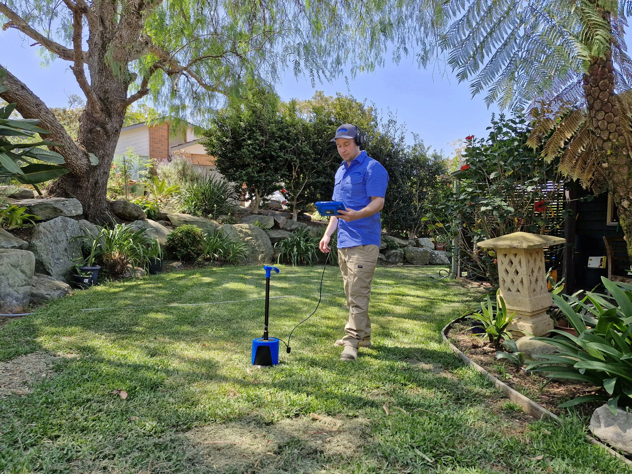 A man wearing a blue shirt, beige pants, and headphones stands in a backyard lawn, operating a metal detector connected to a blue tablet. There are trees, shrubs, and rocks around him, with a house visible in the background.