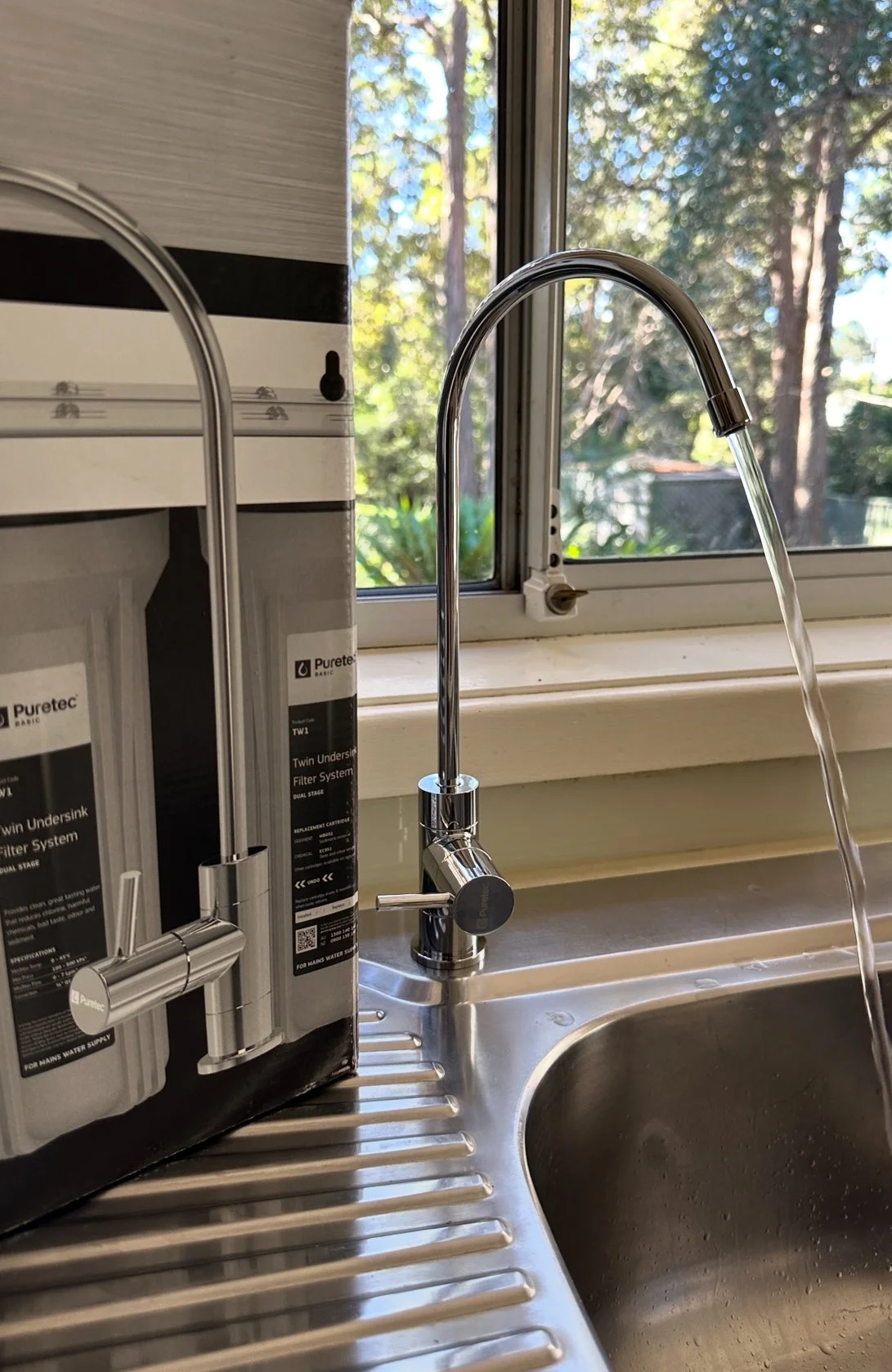 Kitchen sink with a chrome faucet running water, next to a window showing trees outside, and a water filtration system on the left.