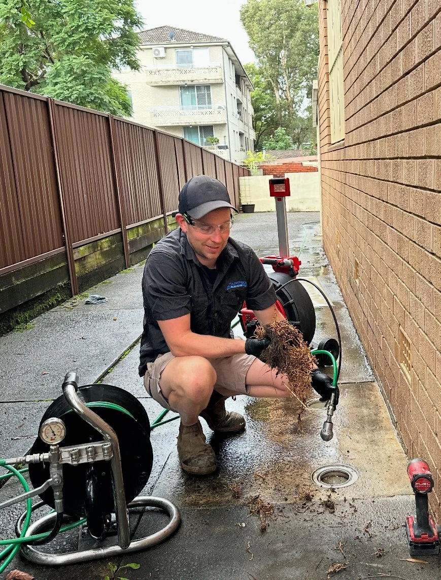 A man kneels on a wet concrete surface holding a clump of dirt and roots, next to a drain at the side of a brick building. He is wearing safety glasses, a cap, a black shirt, and khaki shorts, surrounded by green hoses, a hose reel, and a cordless drill placed on the ground. There is a brown fence and a nearby residential building in the background.
