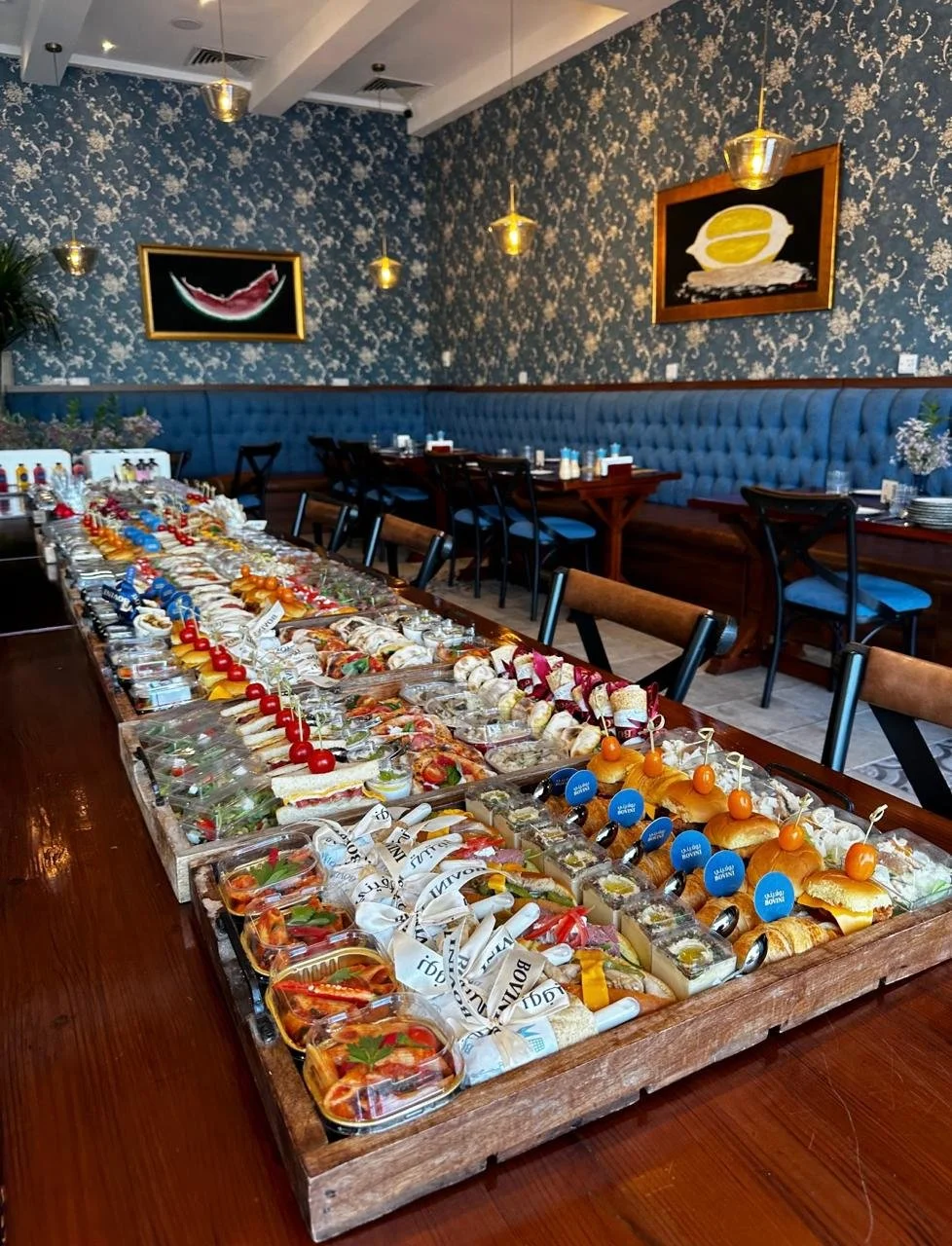 Buffet table filled with assorted appetizers and finger foods in a decorated dining room with blue floral wallpaper and framed artwork.