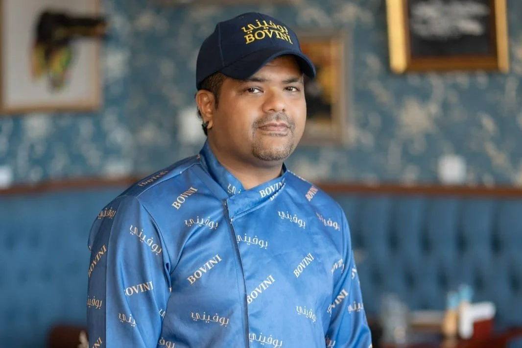 A man wearing a blue shirt and a navy blue cap with 'BOVINI' written on it, standing inside a restaurant with framed artwork on the wall in the background.