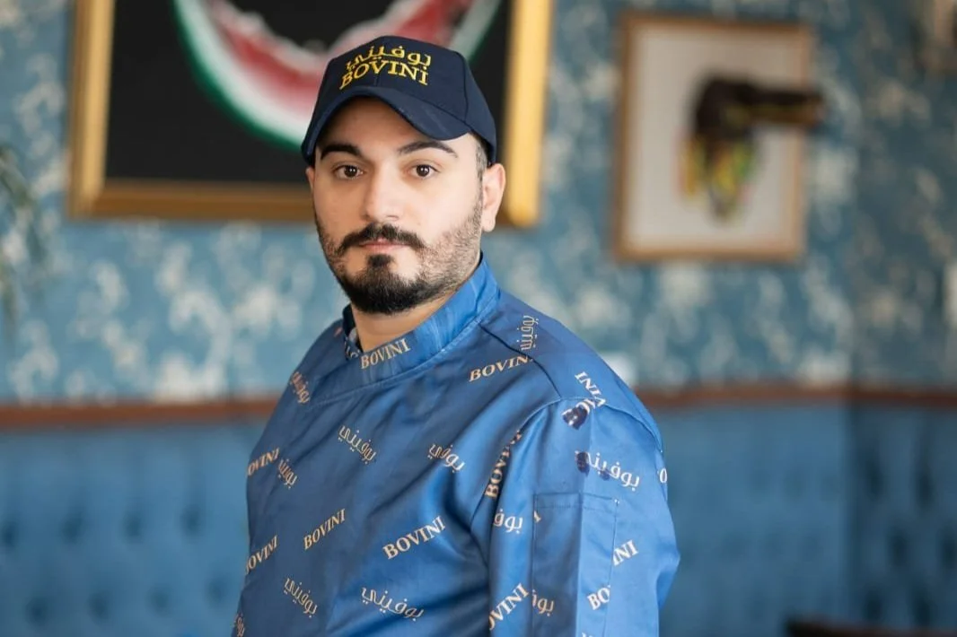 Man in a blue BOVINI branded chef uniform and navy cap looking at the camera with a blurred background of framed pictures on a blue patterned wall.