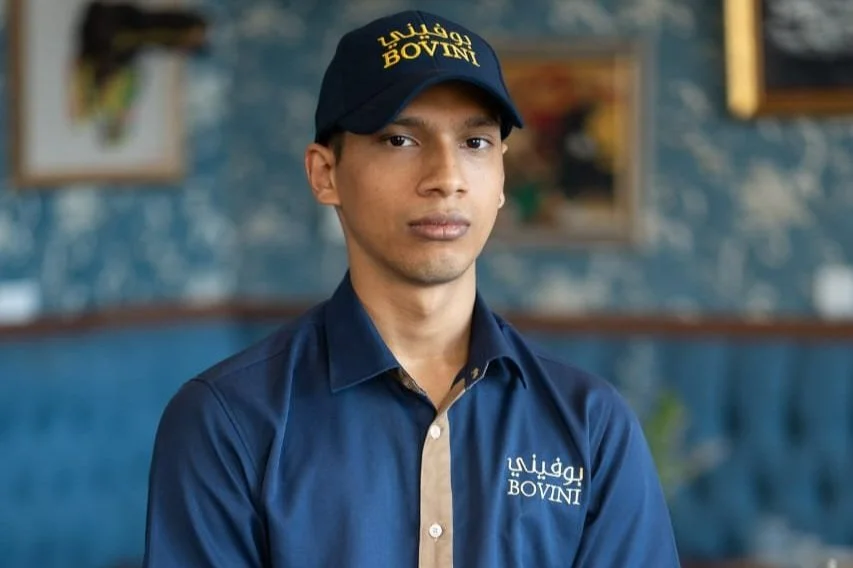 Young man wearing a navy blue cap and matching shirt with 'BOVIN' logo, standing indoors with a blurred blue patterned wall and framed pictures in the background.